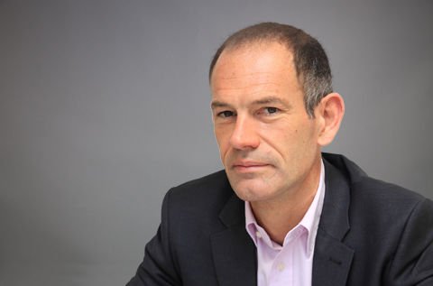 A man with short brown hair, dressed in a black blazer and light pink shirt, poses against a plain gray background.