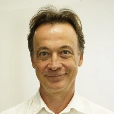 Portrait of a man with dark hair smiling at the camera, wearing a white shirt, against a plain light-colored background.