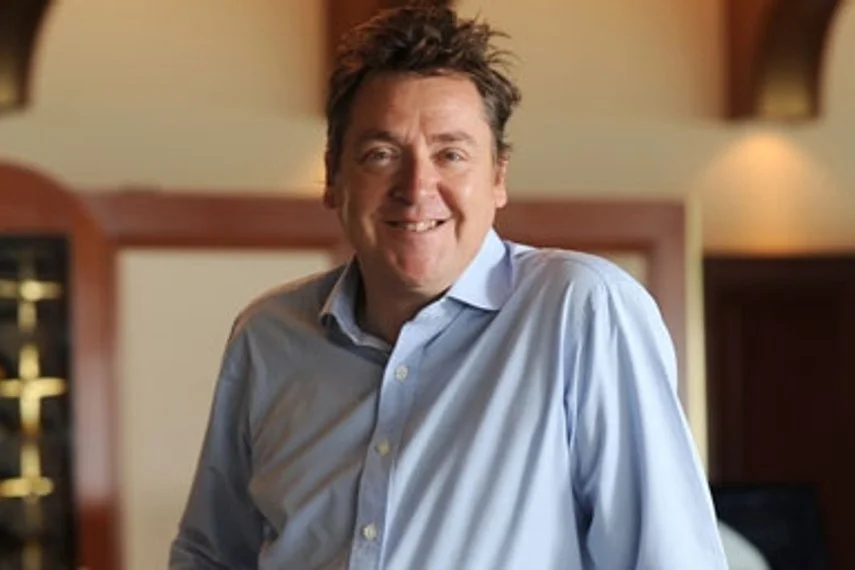 A man with dark, slightly curly hair, wearing a light blue dress shirt, smiling, indoors in a warmly lit room with wooden shelves or cabinets in the background.