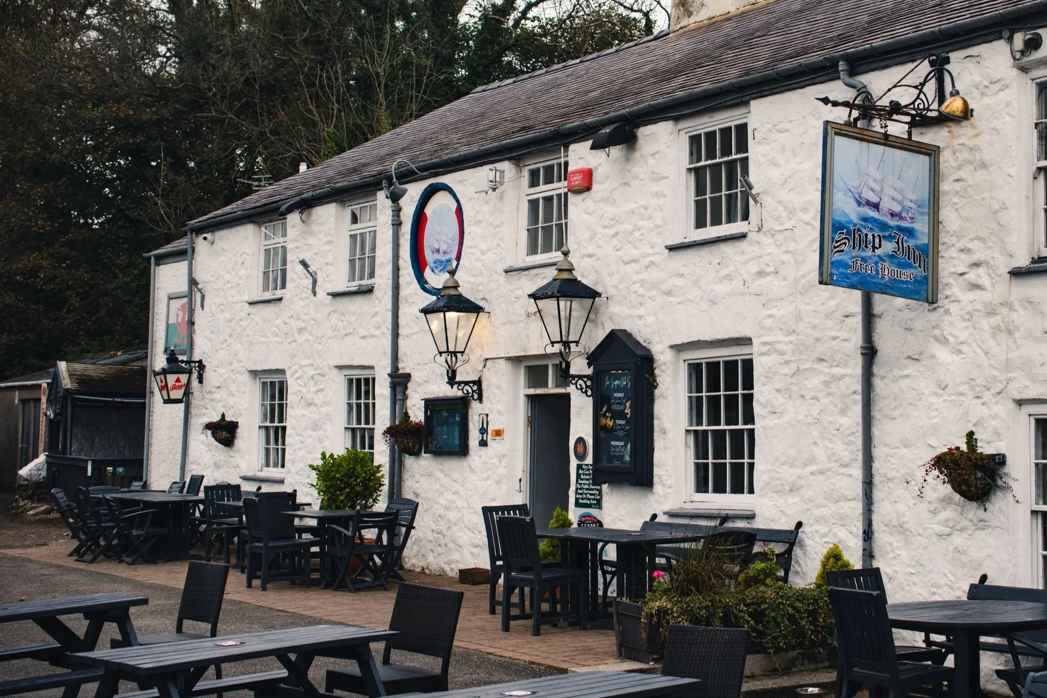 18th-century white stone pub exterior and historic 'Ship Inn Free House' sign, featuring a dog-friendly beer garden, black tables and chairs, vintage lamps, and plants. The authentic inn is located directly on the beach and the Anglesey Coastal Path