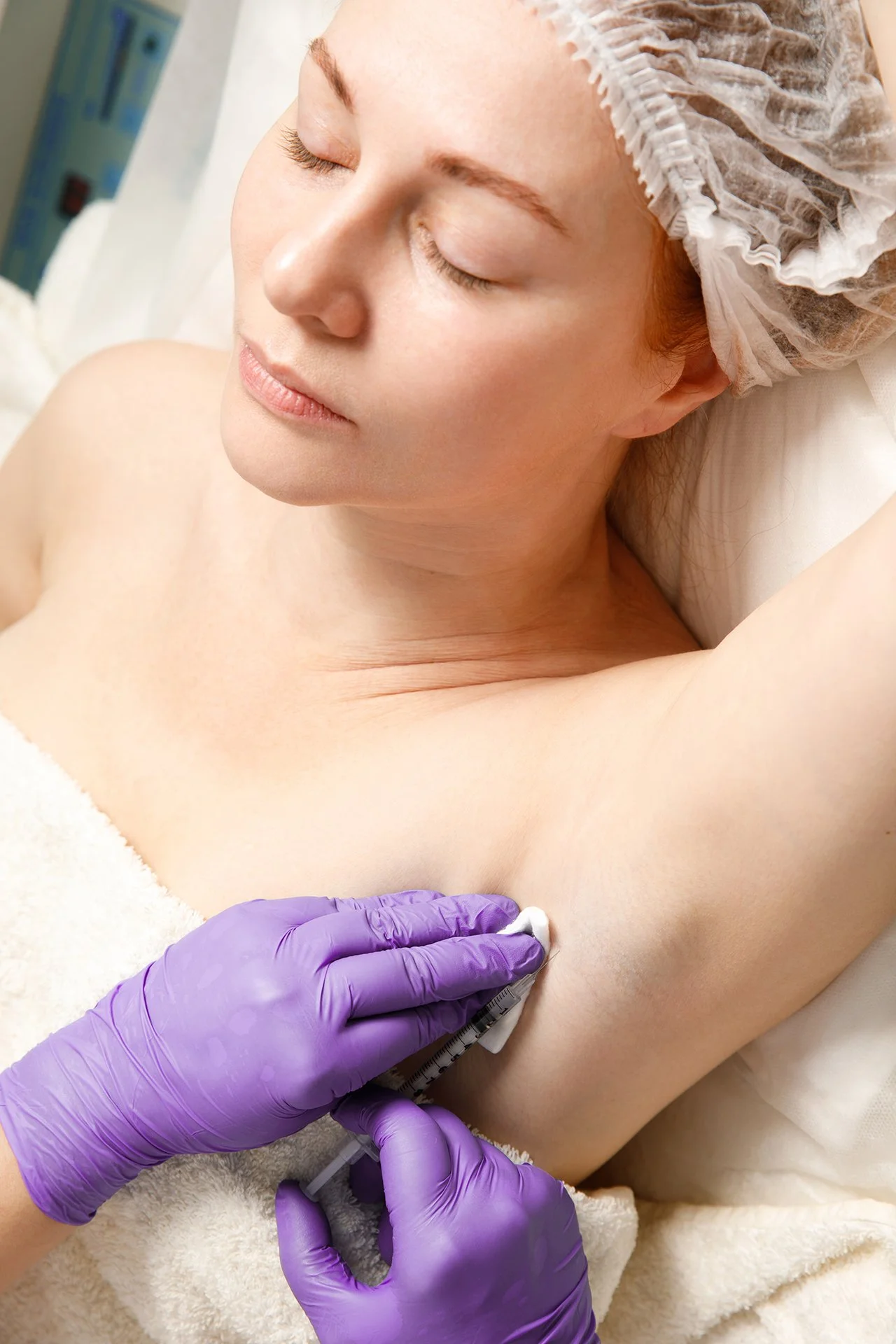 A woman receiving an injection under her left arm while lying down with her eyes closed. She is wearing a disposable cap, and a healthcare professional wearing purple gloves is administering the injection and wiping the area.