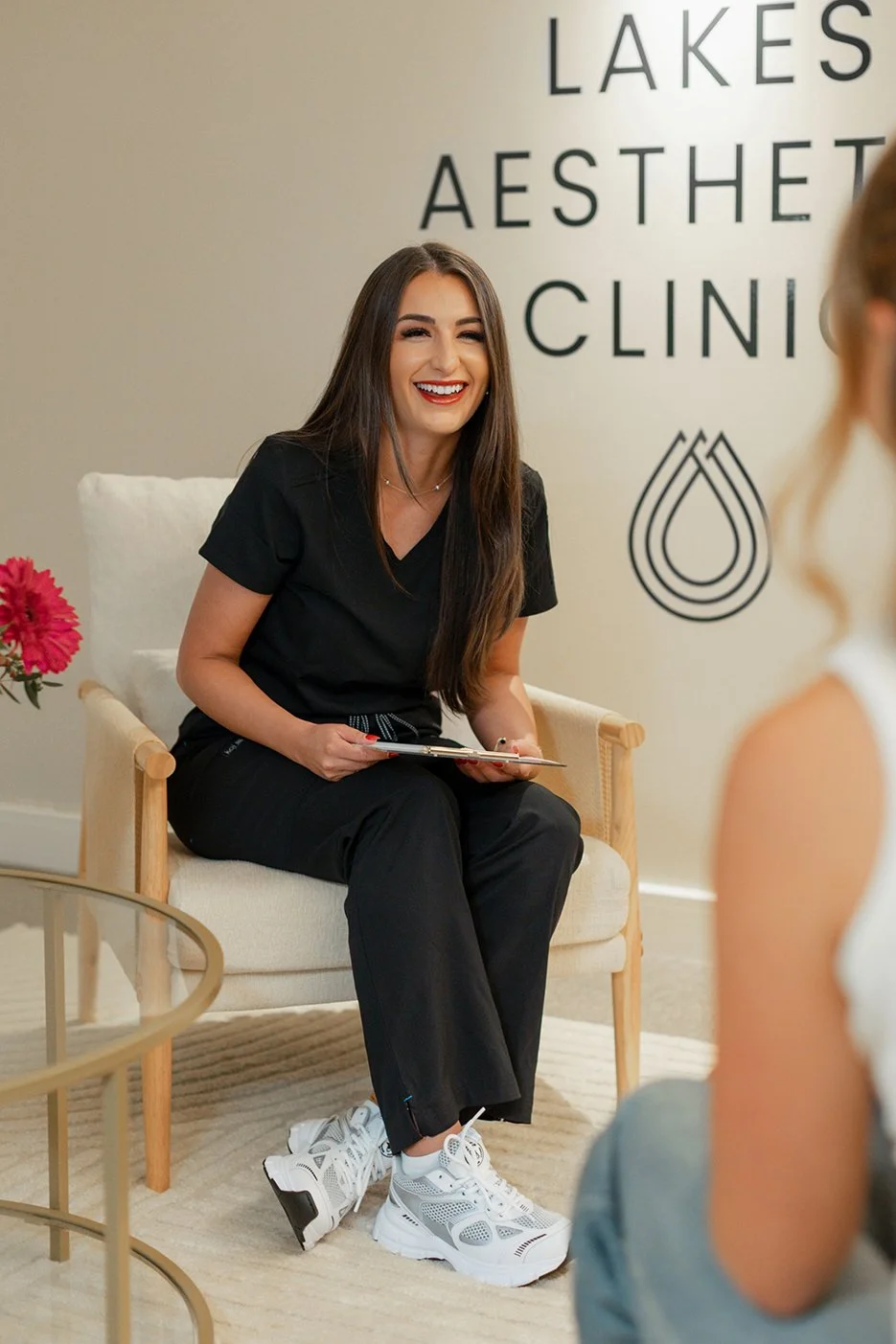 A woman in black scrubs sitting on a chair at a skincare or aesthetic clinic, talking to a client. The clinic's name is partially visible on the wall behind her, reading 'LAKES AESTHETIC CLINIC', with a water droplet logo next to it.