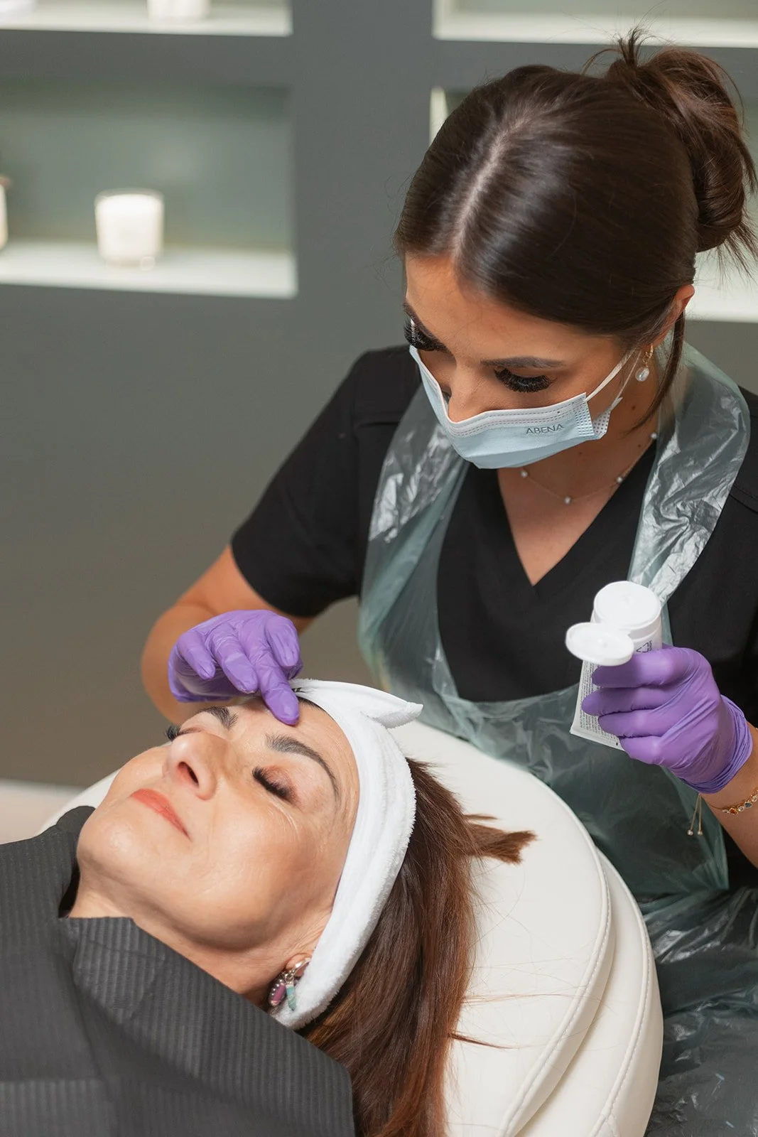 A woman receiving a facial treatment from an aesthetician wearing gloves, a mask, and a plastic apron.