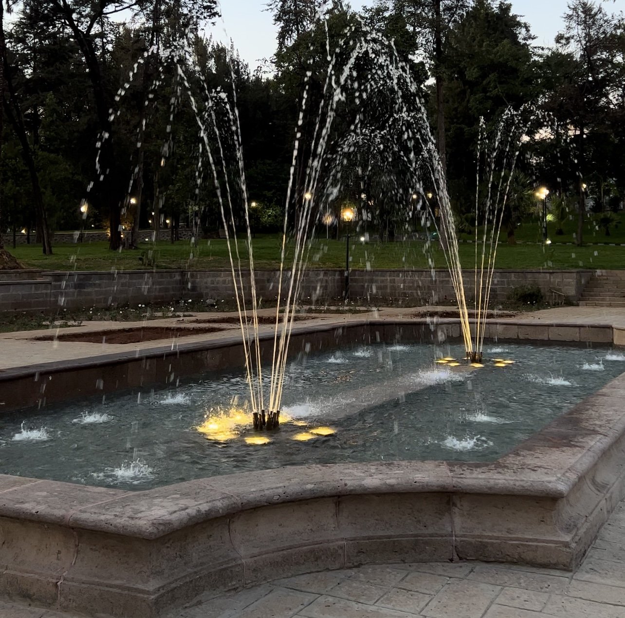 A stone fountain with multiple water jets illuminated by yellow lights, in a park at dusk or evening, with trees and park lights in the background.