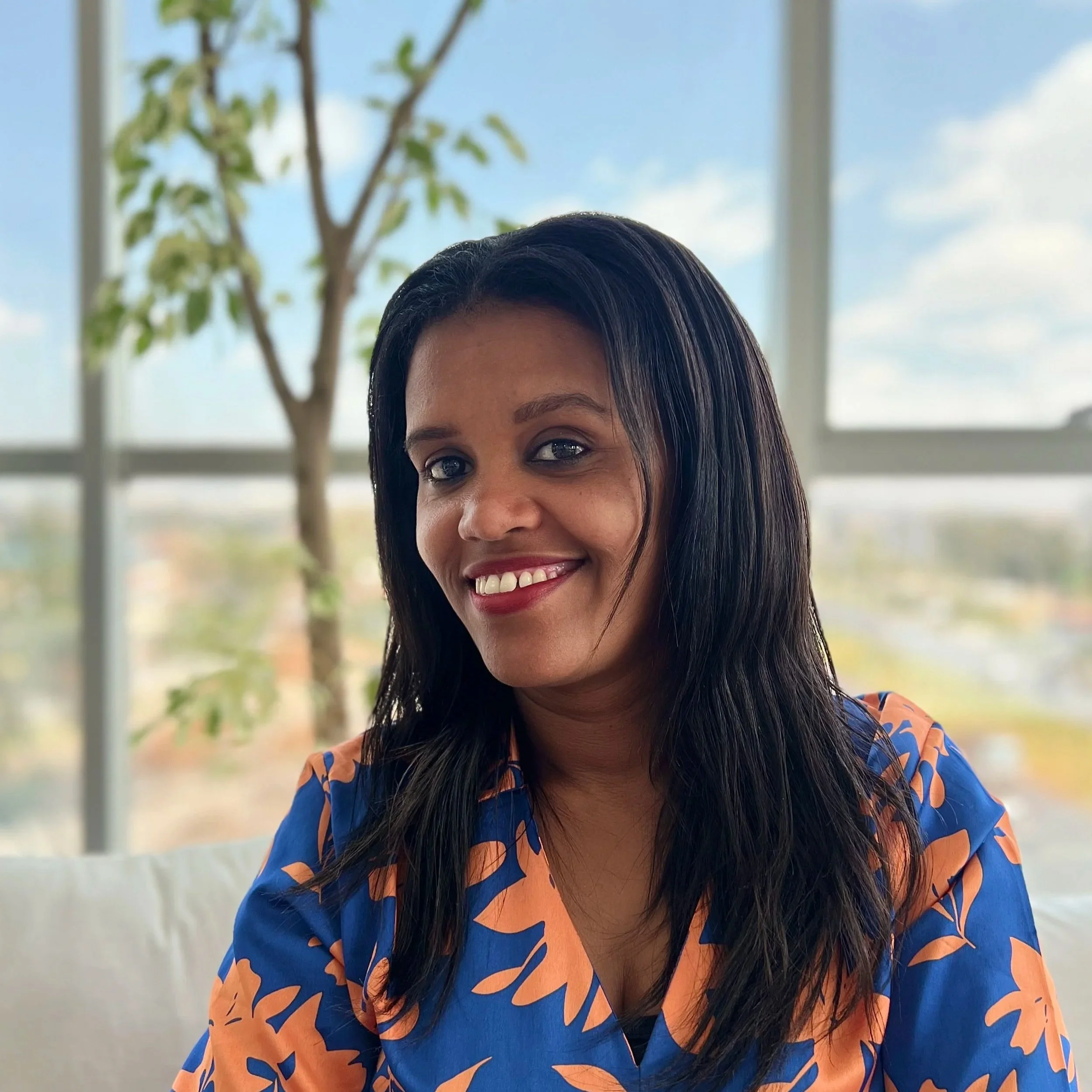 A woman with long black hair and a bright smile seated in a room with large windows showing a blue sky and a tree in the background, wearing a colorful floral top.