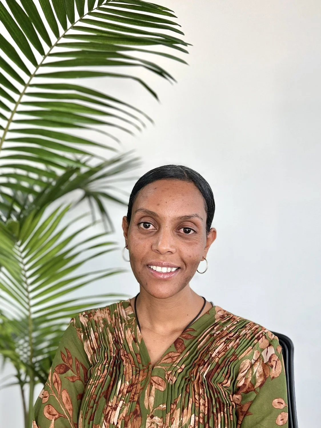 A young woman with dark hair pulled back, wearing hoop earrings, a green floral blouse, sits in front of a white wall and a large green palm plant.