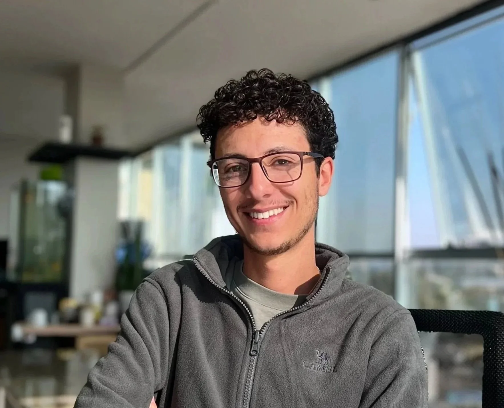 Young man with curly dark hair, glasses, and a gray zip-up hoodie smiling indoors near large windows.