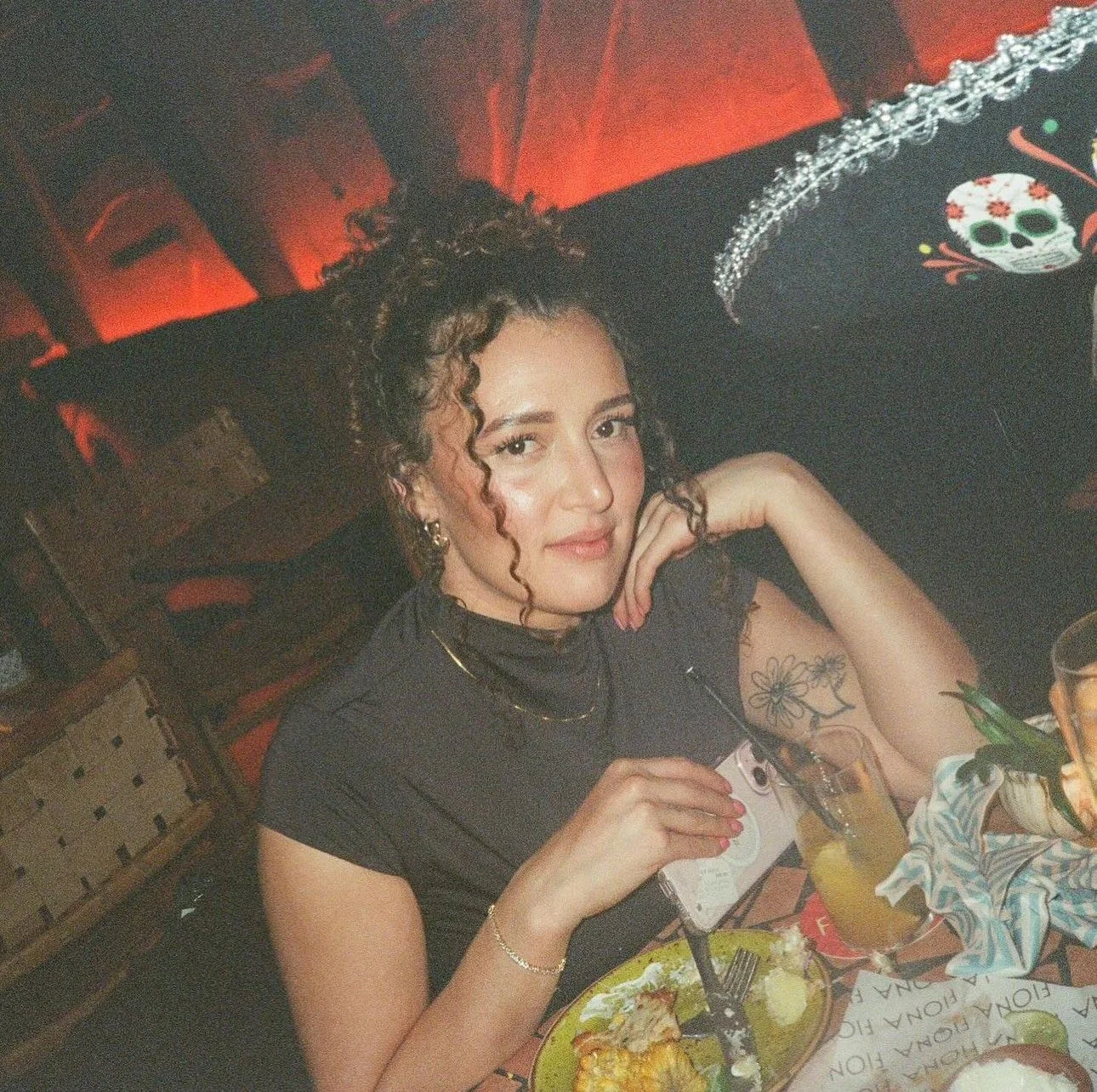 A woman with curly hair and tattoos is sitting at a table in a restaurant, holding a fork and a camera, with a plate of food and drinks on the table, and decorative Día de los Muertos masks in the background.