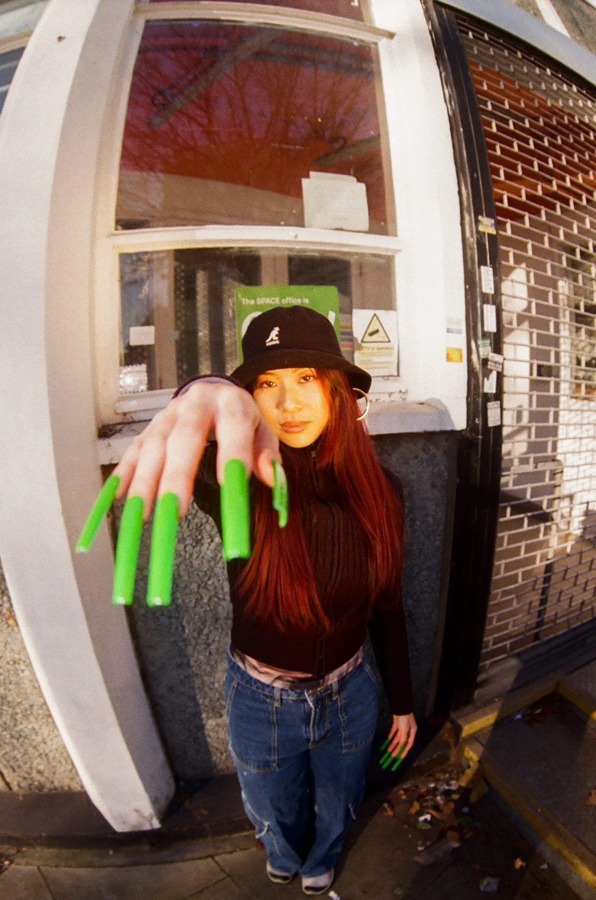 Young woman with long red hair wearing a black bucket hat, black zip-up top, and baggy jeans, standing outside in front of a brick wall and window, reaching out towards the camera with her hand featuring long neon green nails.
