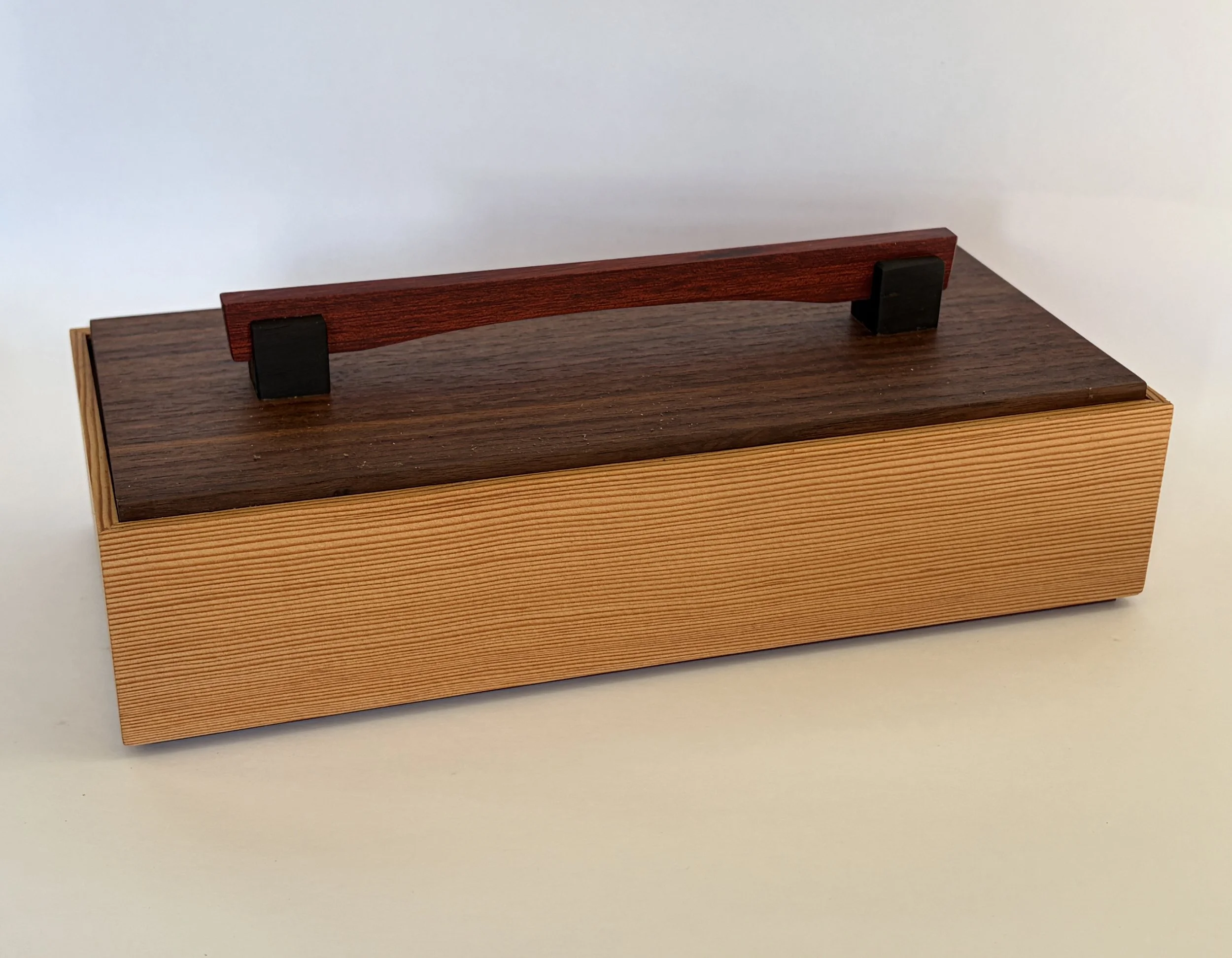 A wooden box with a dark brown interior and a lighter wood exterior. On top of the box, there is a small, curved, reddish-brown wooden handle supported by two small black blocks.