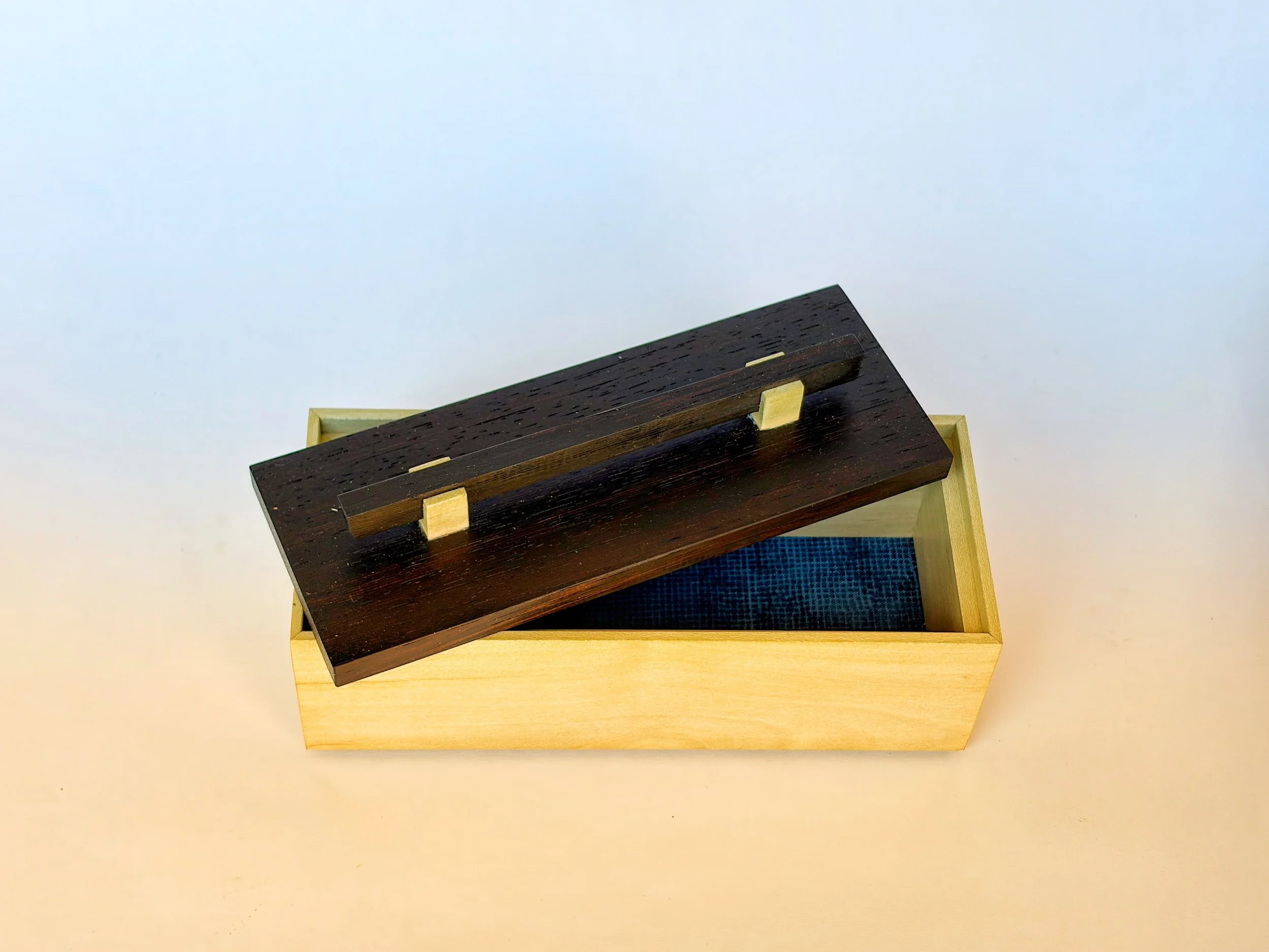 A box made of holly.  The dark wood lid fits just inside the box.  The bottom of the box is lined with blue fabric.