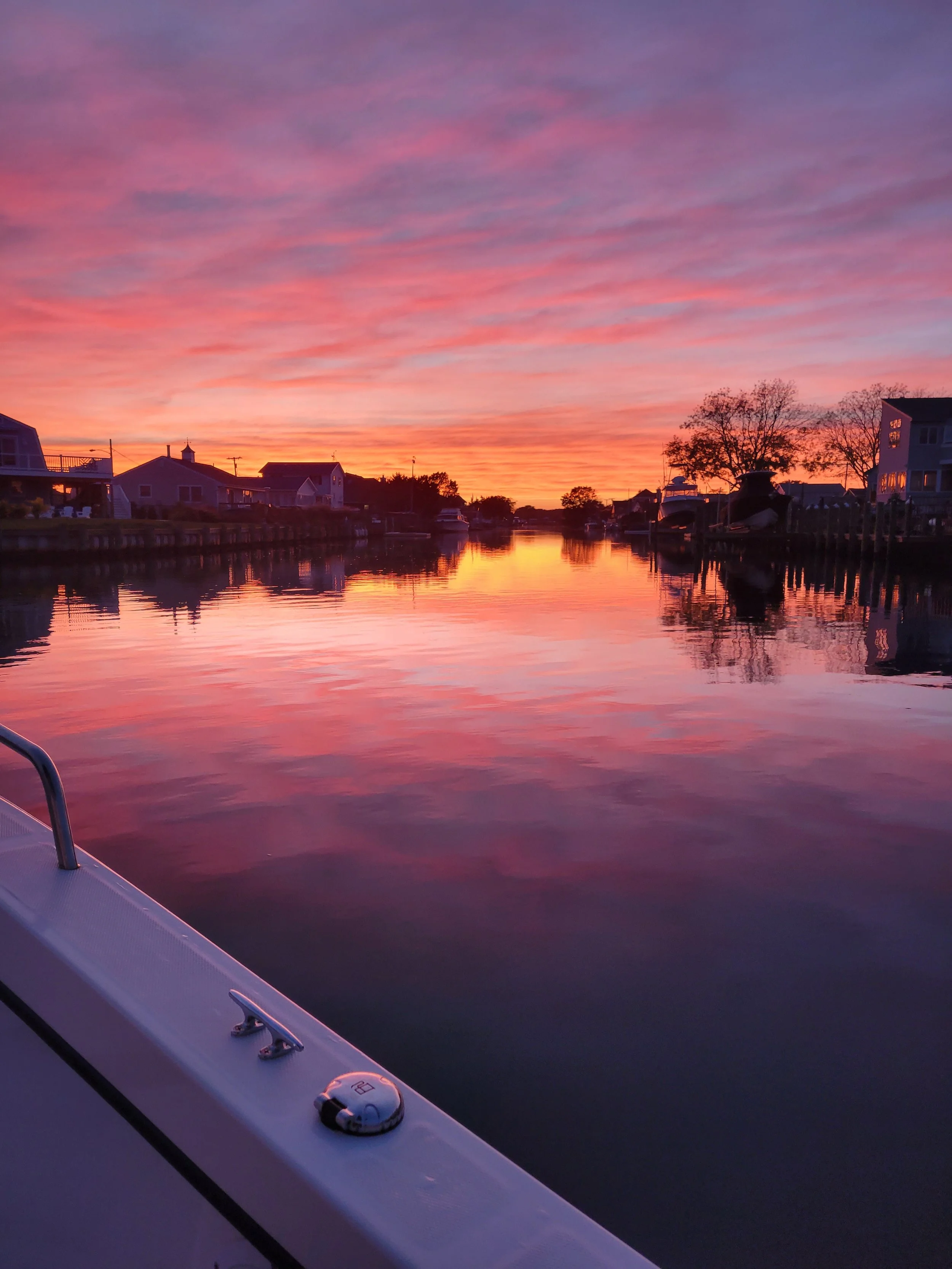 Colorful sunset over a calm waterway with boats and houses on both sides, reflected in the water, seen from a boat.