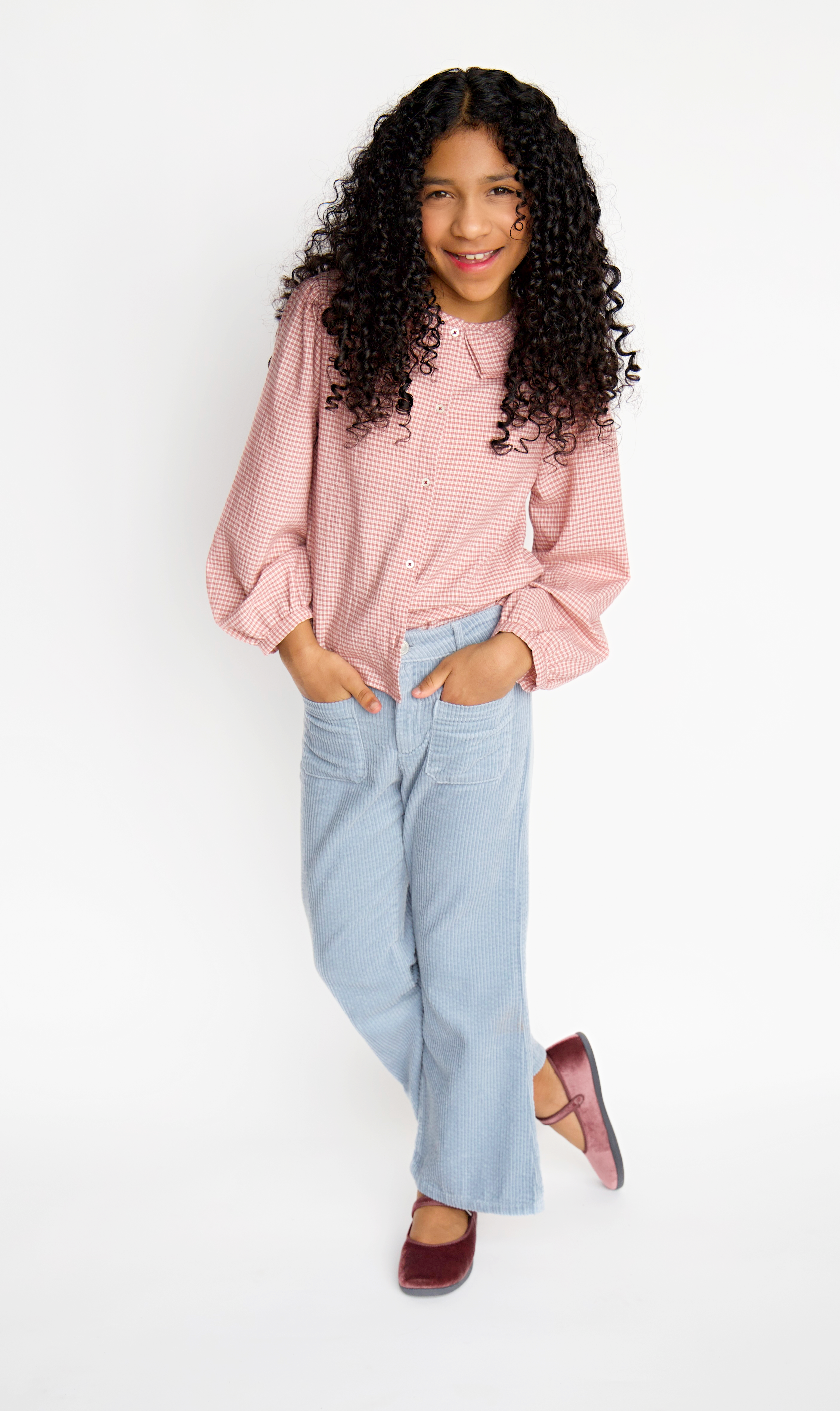 A young girl with curly hair, wearing a pink checkered long-sleeve shirt, light blue pants, and burgundy shoes, standing against a white background.