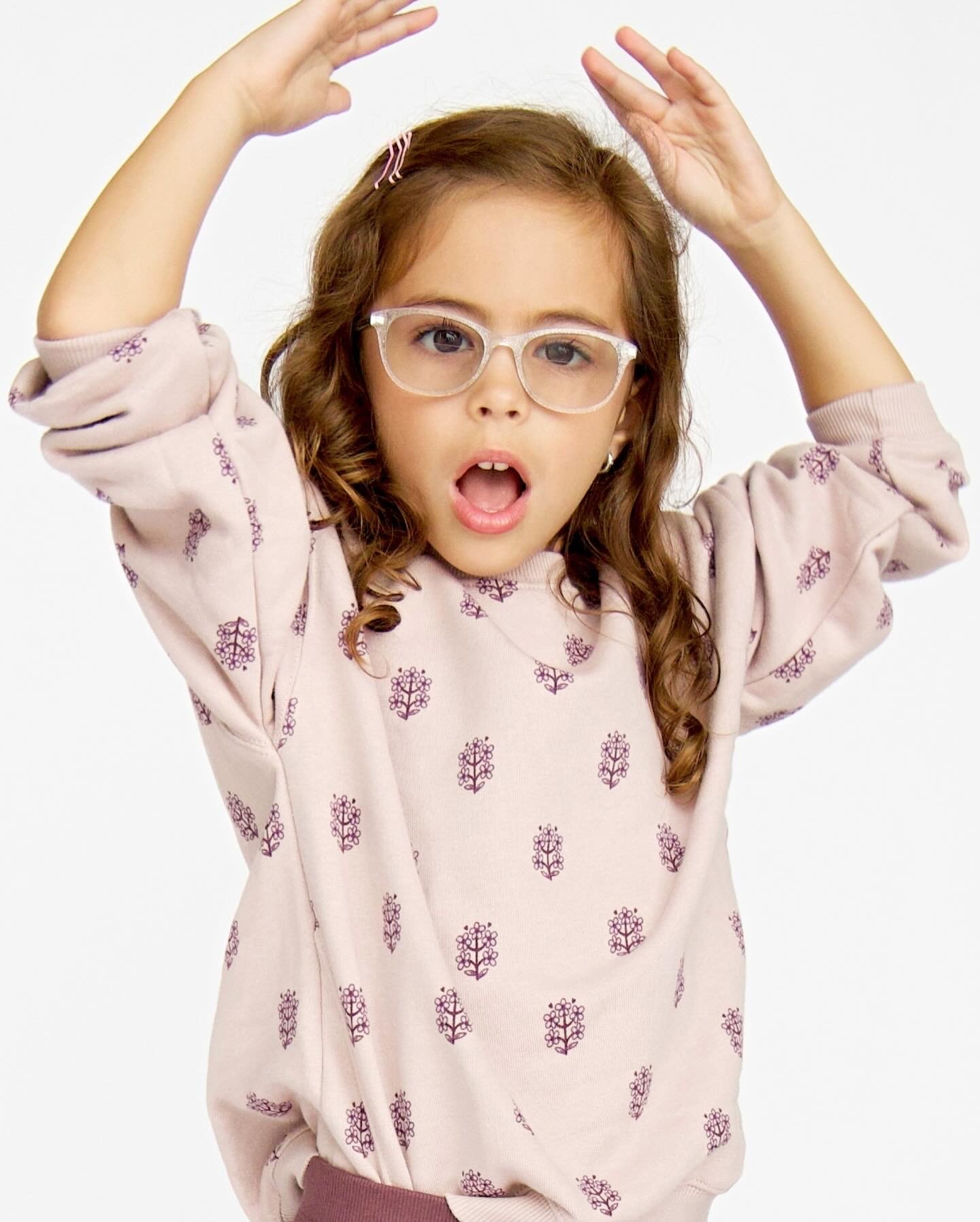 A young girl with curly brown hair, wearing clear glasses and a beige sweatshirt with purple floral patterns, looks surprised with her mouth open, raising her hands above her head.