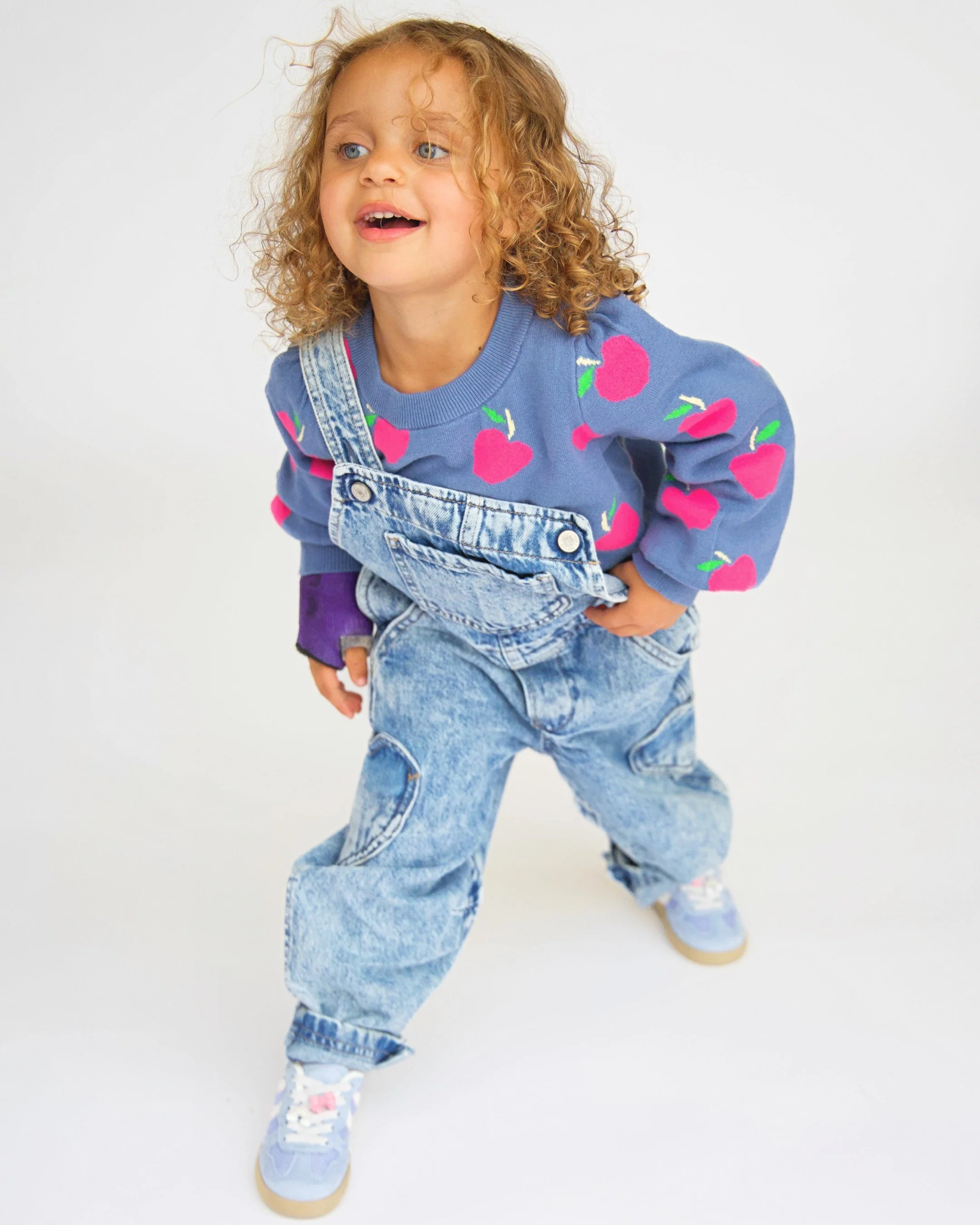 A young girl with curly blonde hair and blue eyes wearing a blue sweater with pink apples, overalls, and sneakers, smiling and leaning forward against a white background.