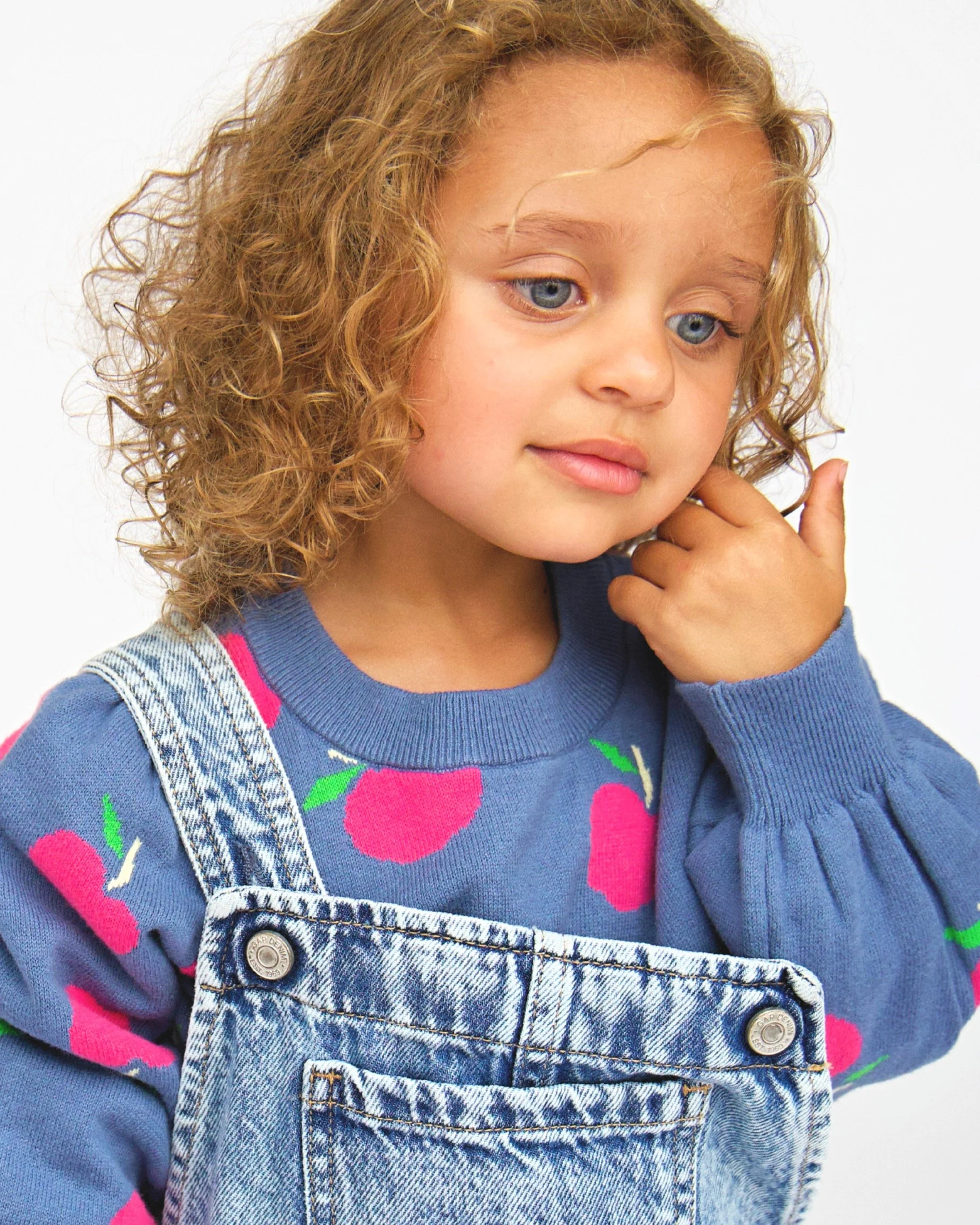Young girl with curly blonde hair and blue eyes wearing a blue sweater with pink cherry designs and denim overalls, touching her face with her hand.