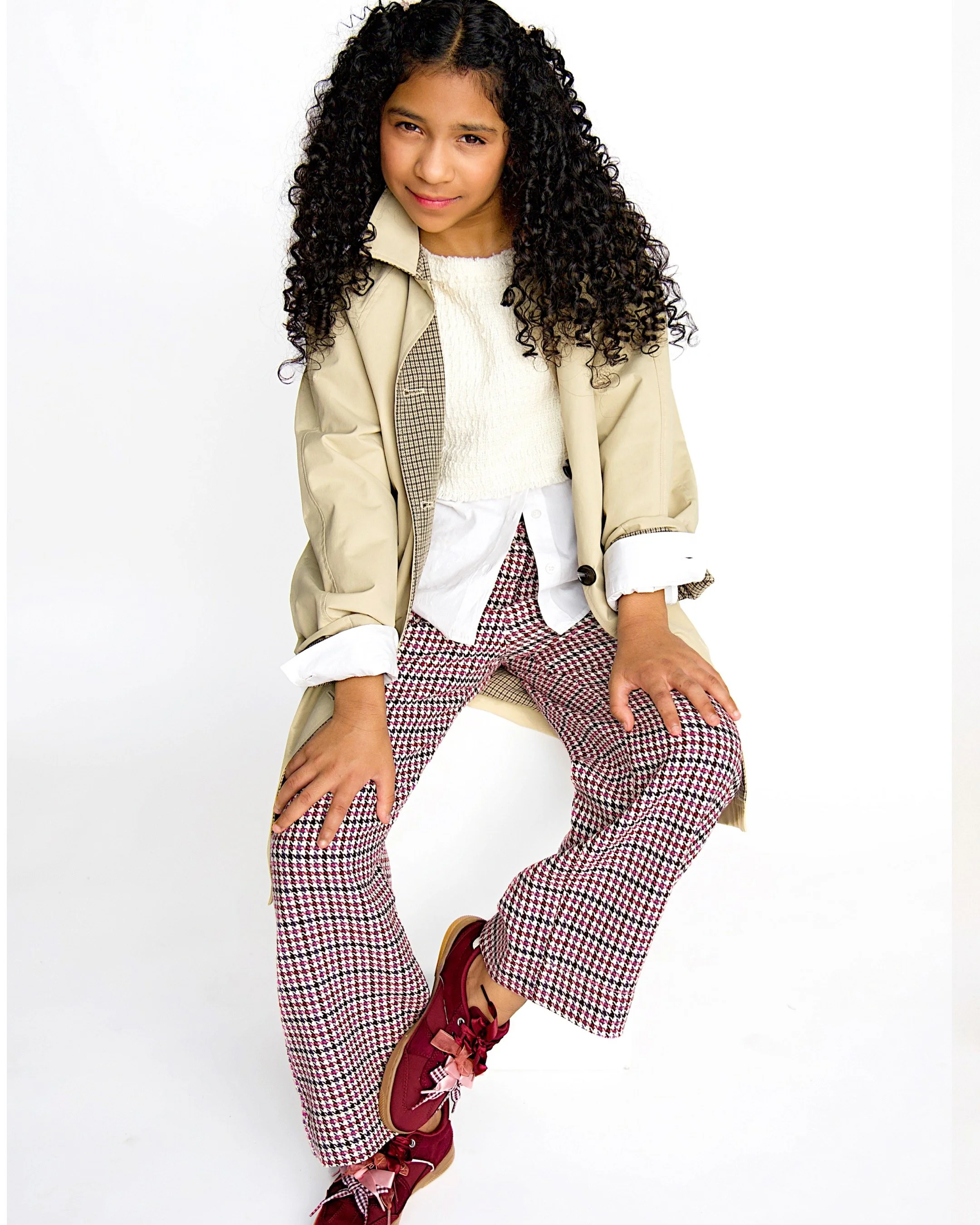 A young girl with curly hair wearing a beige coat, a white sweater, plaid pants, and red sneakers, posing against a plain white background.