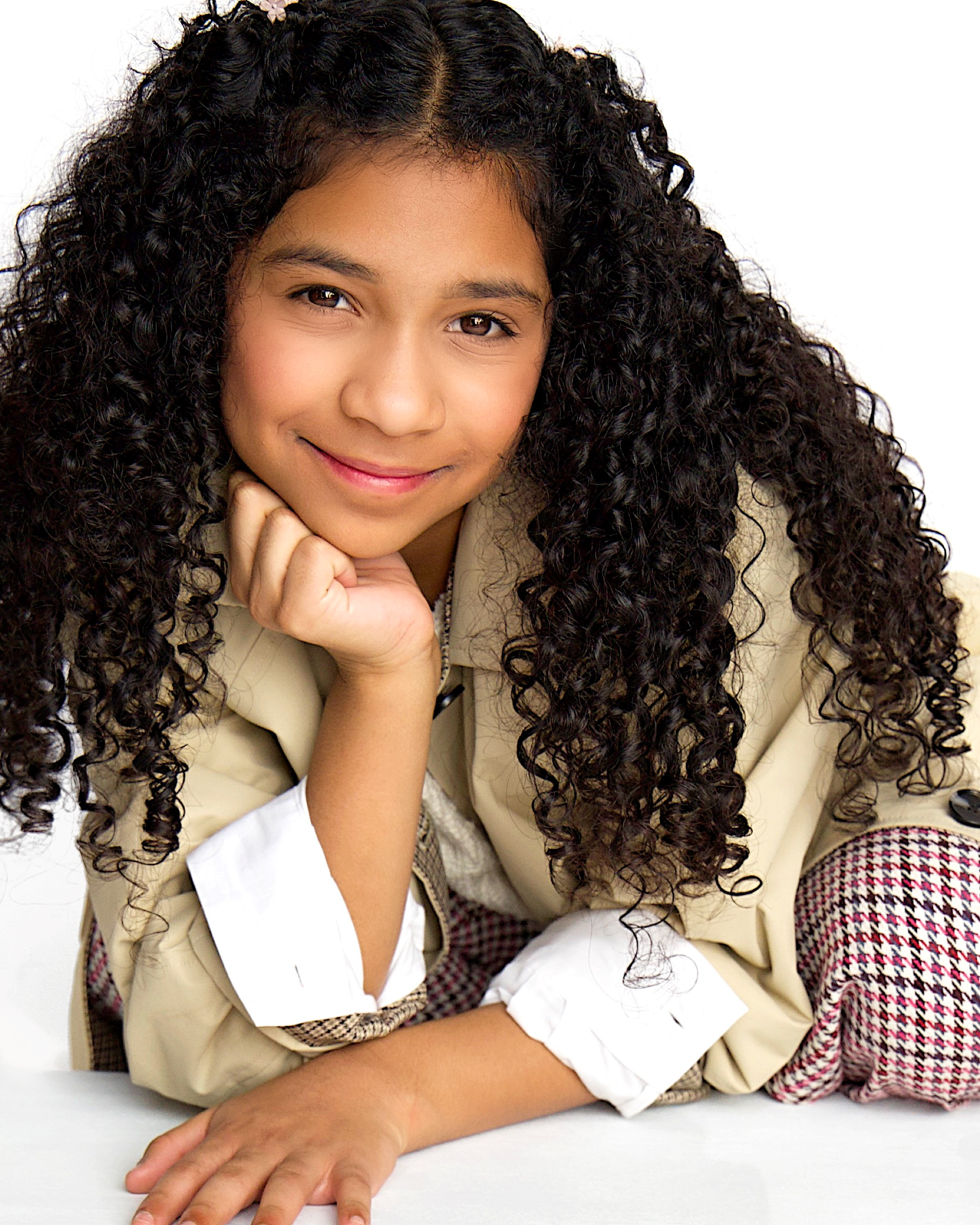 A young girl with long, curly hair smiling and resting her chin on her hand, wearing a beige jacket and white shirt, lying on a white surface.
