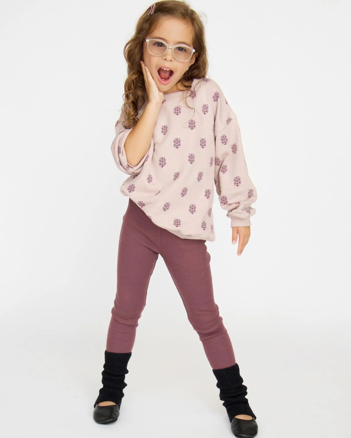 A young girl with curly hair, wearing glasses, a pink sweater with purple patterns, maroon pants, black leg warmers, and black shoes, posing with her mouth open and one hand on her face.