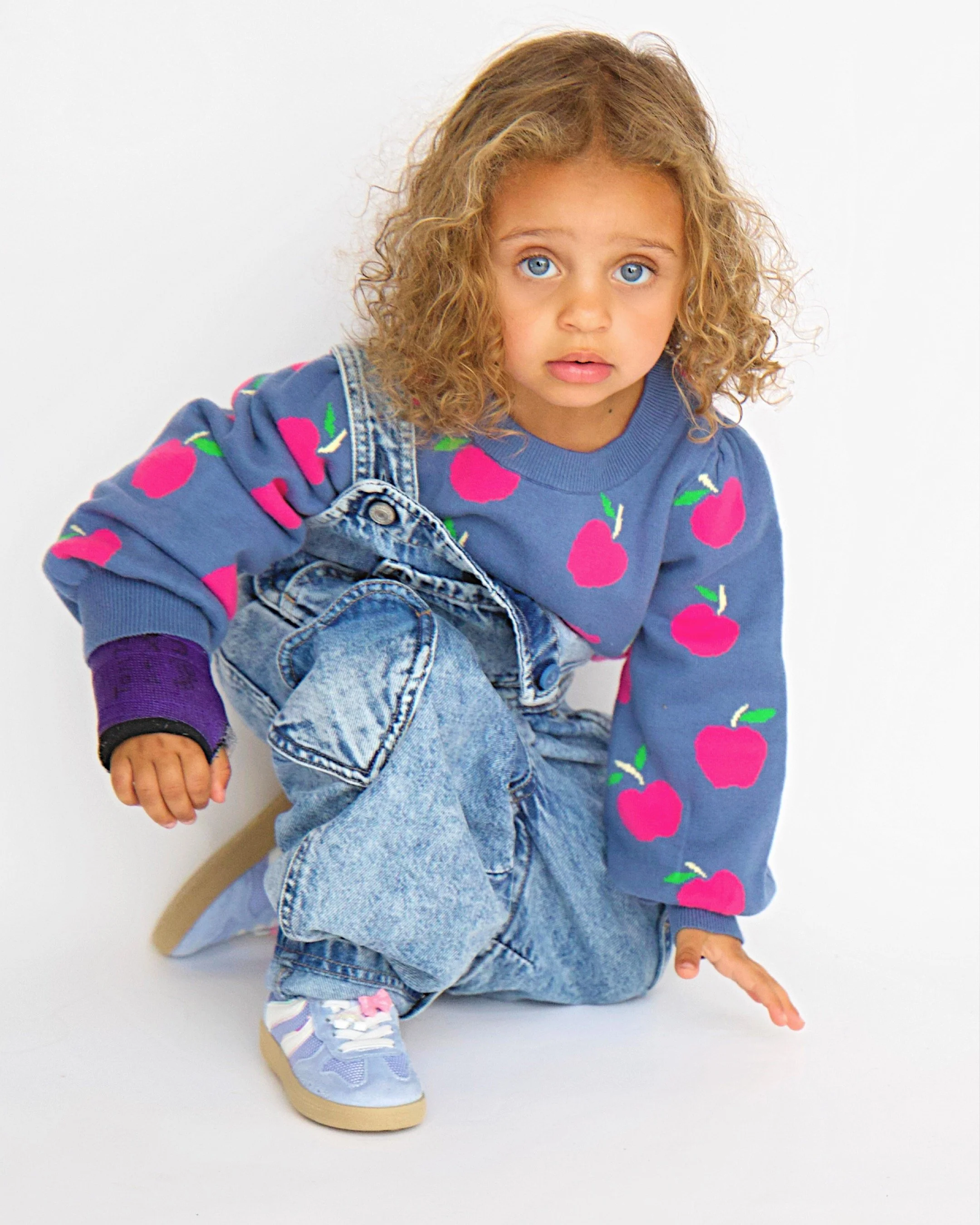 Young girl with curly hair and blue eyes crouching against a plain background, wearing a blue sweater with pink apple patterns, oversized denim jeans, and pastel sneakers.