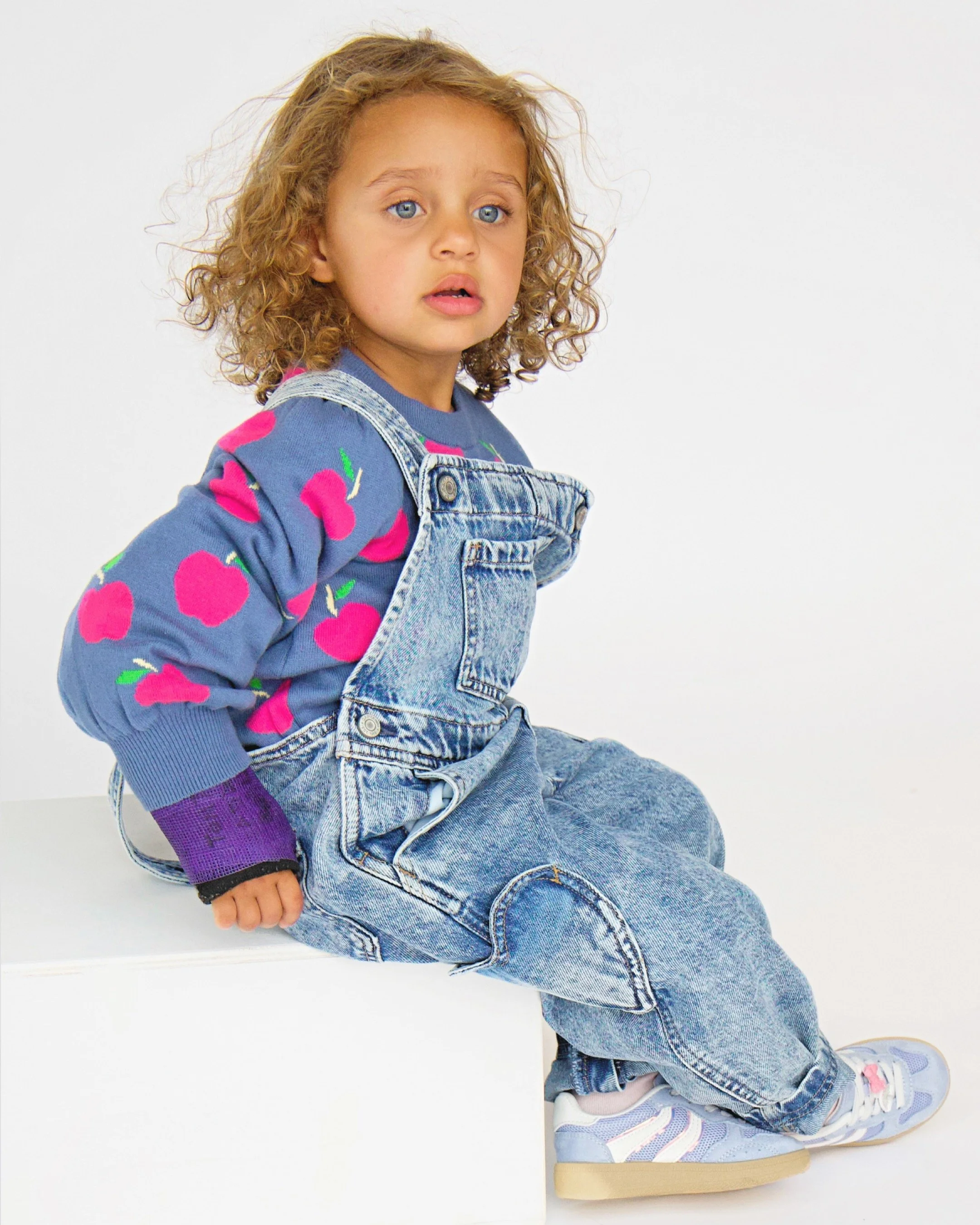 A young girl with curly brown hair and blue eyes sitting against a plain white background, wearing a colorful sweater with pink cherries, denim overalls, and multicolored sneakers, looking thoughtful.
