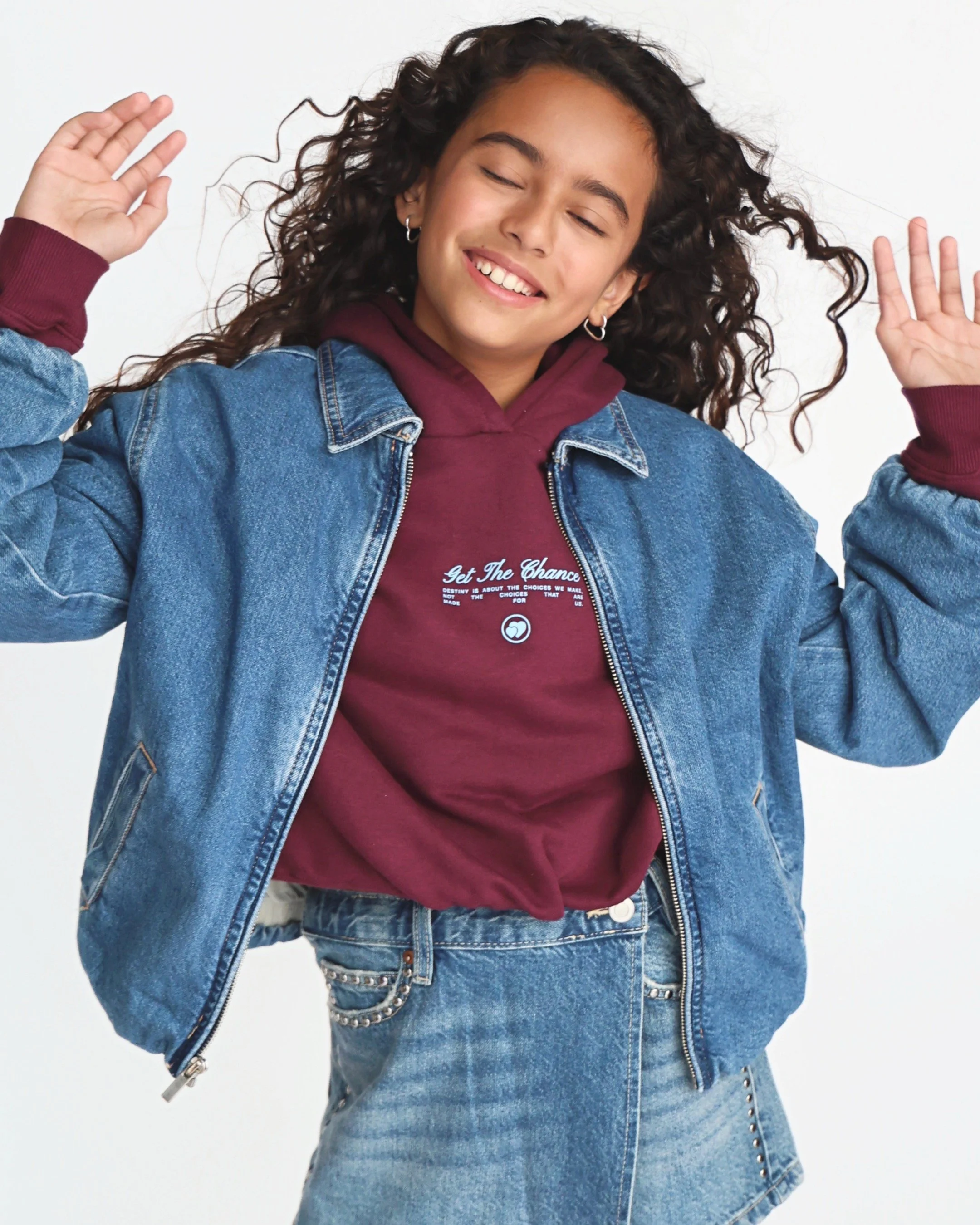Young woman with curly hair smiling with eyes closed, wearing a denim jacket over a maroon hoodie, and light-wash denim shorts, standing against a white background.