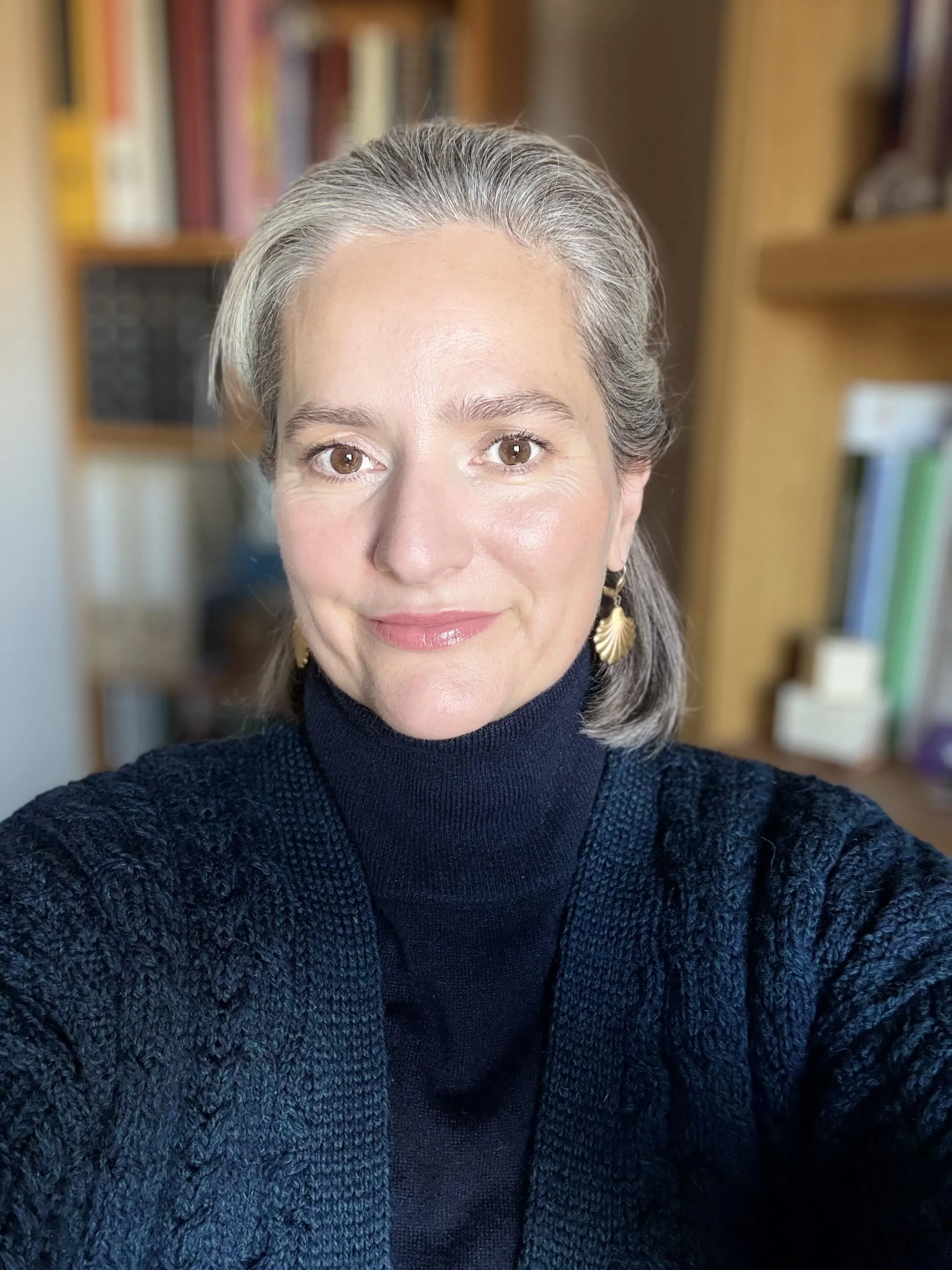 A woman taking a selfie with brown eyes, gray hair, and earrings shaped like seashells, wearing a black turtleneck and a dark knitted cardigan, in a room with wooden bookshelves filled with books.