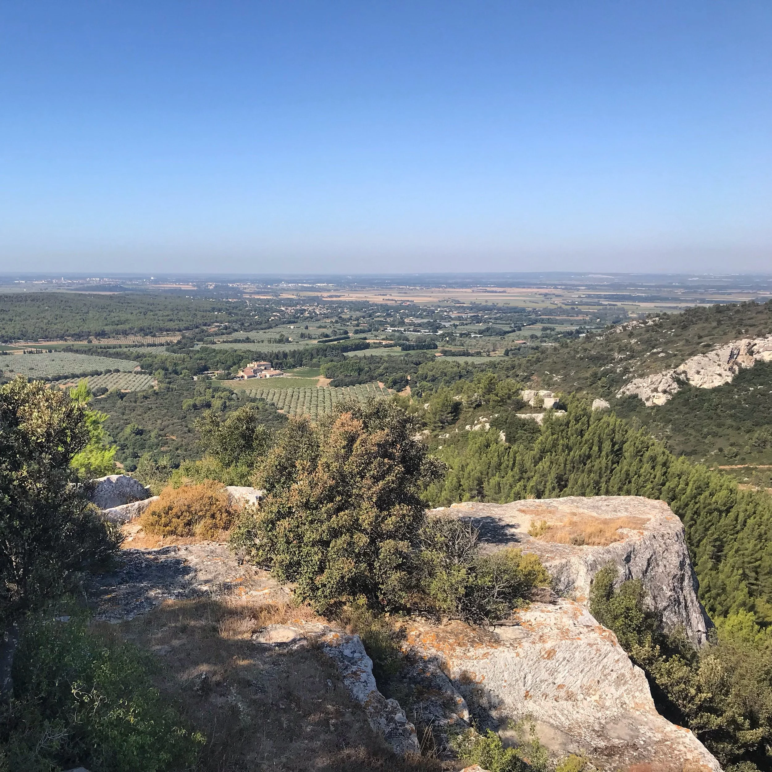 A scenic view from a rocky hillside overlooking a lush green valley with trees, farmland, and distant hills under a clear blue sky.