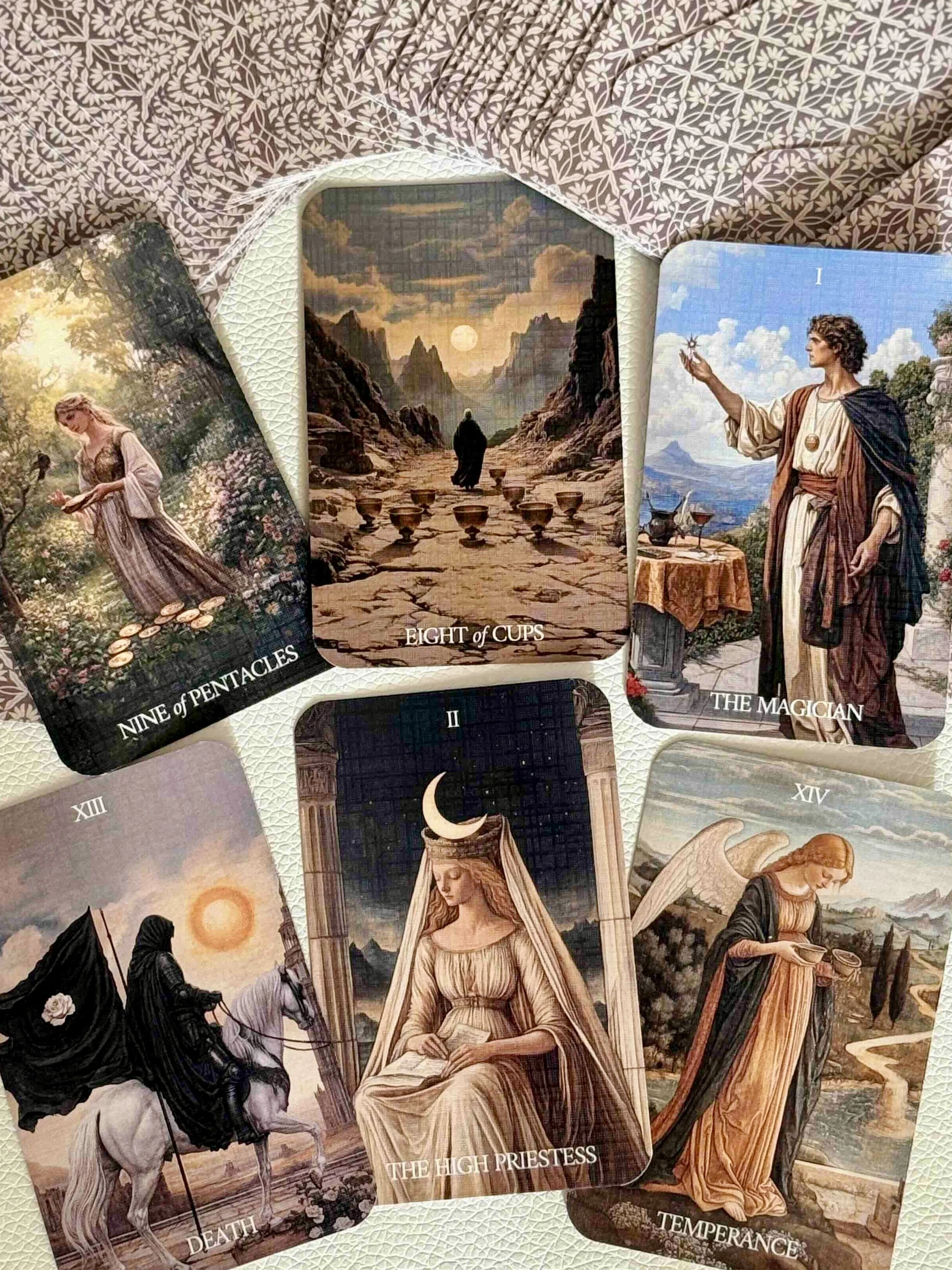 A spread of six tarot cards including the Nine of Pentacles, Eight of Cups, The Magician, Death, The High Priestess, and Temperance, displayed on a white textured surface with a floral pattern.