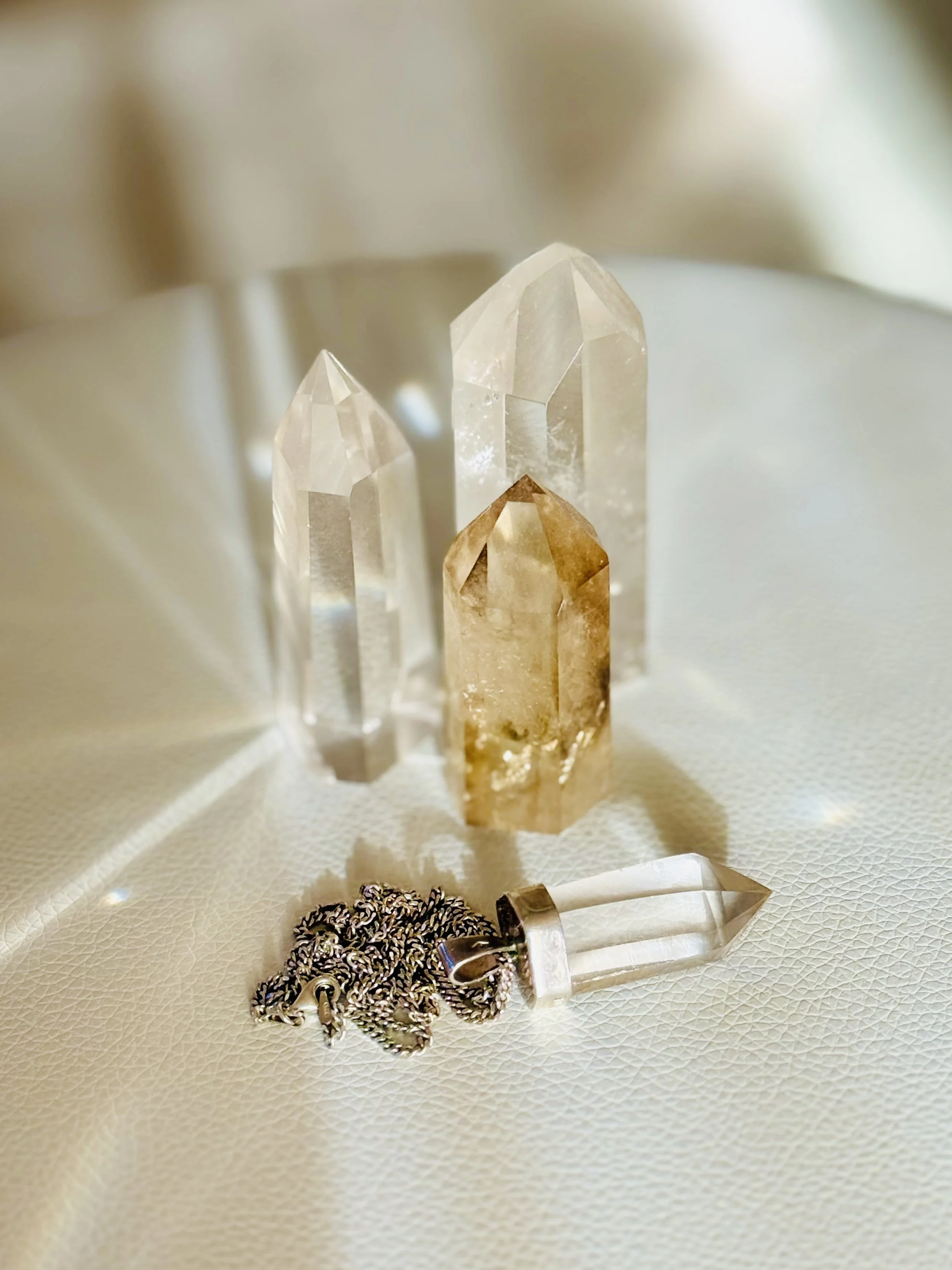 Three large geode crystals, two clear and one brownish, along with a small clear pendant crystal with silver chain, all placed on a textured ivory surface.