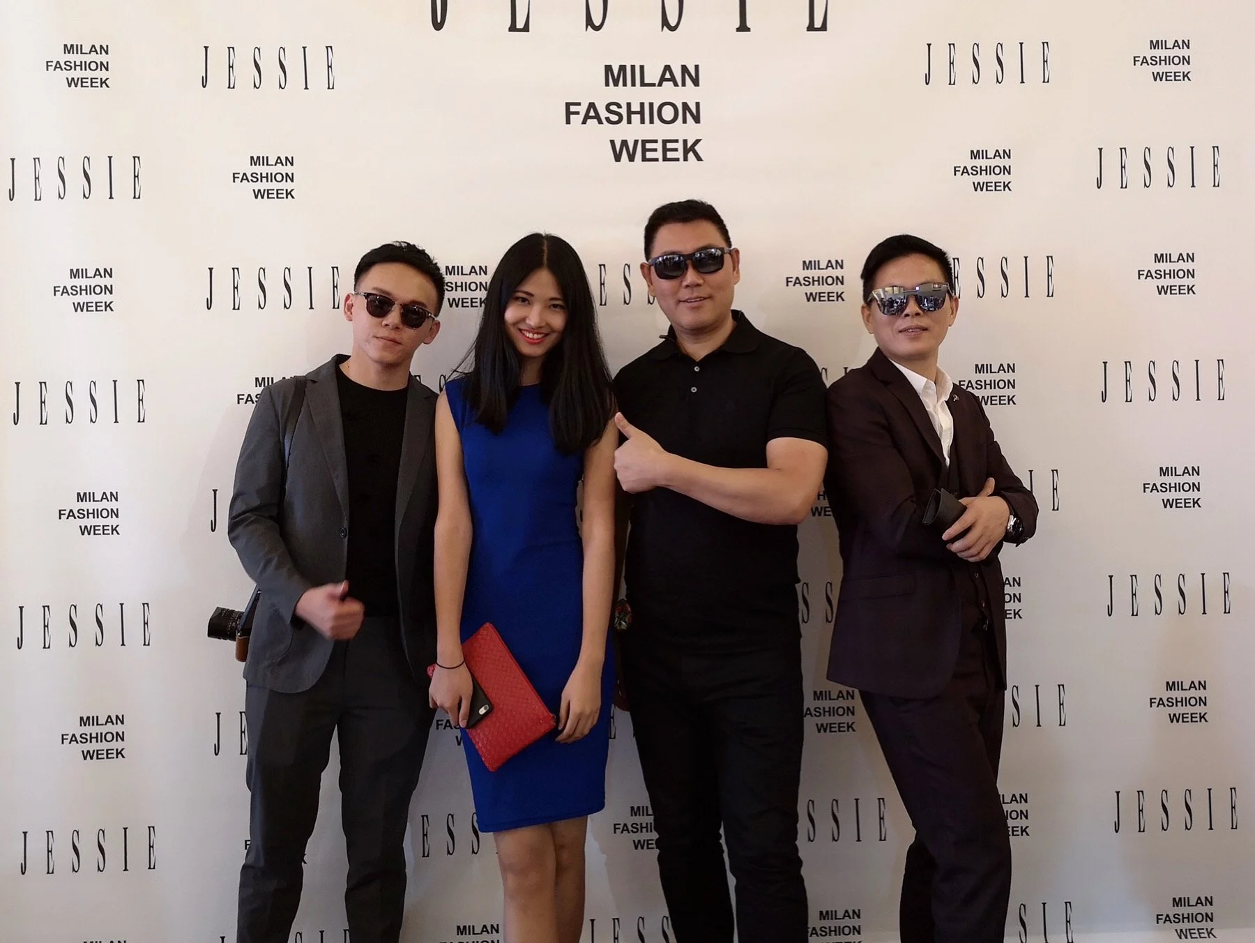 Four people standing in front of a backdrop that reads "Milan Fashion Week" and "Jessie." The group includes two men with sunglasses, a woman in a royal blue dress holding a red clutch, and a man in a black shirt. They are posing together at a fashio