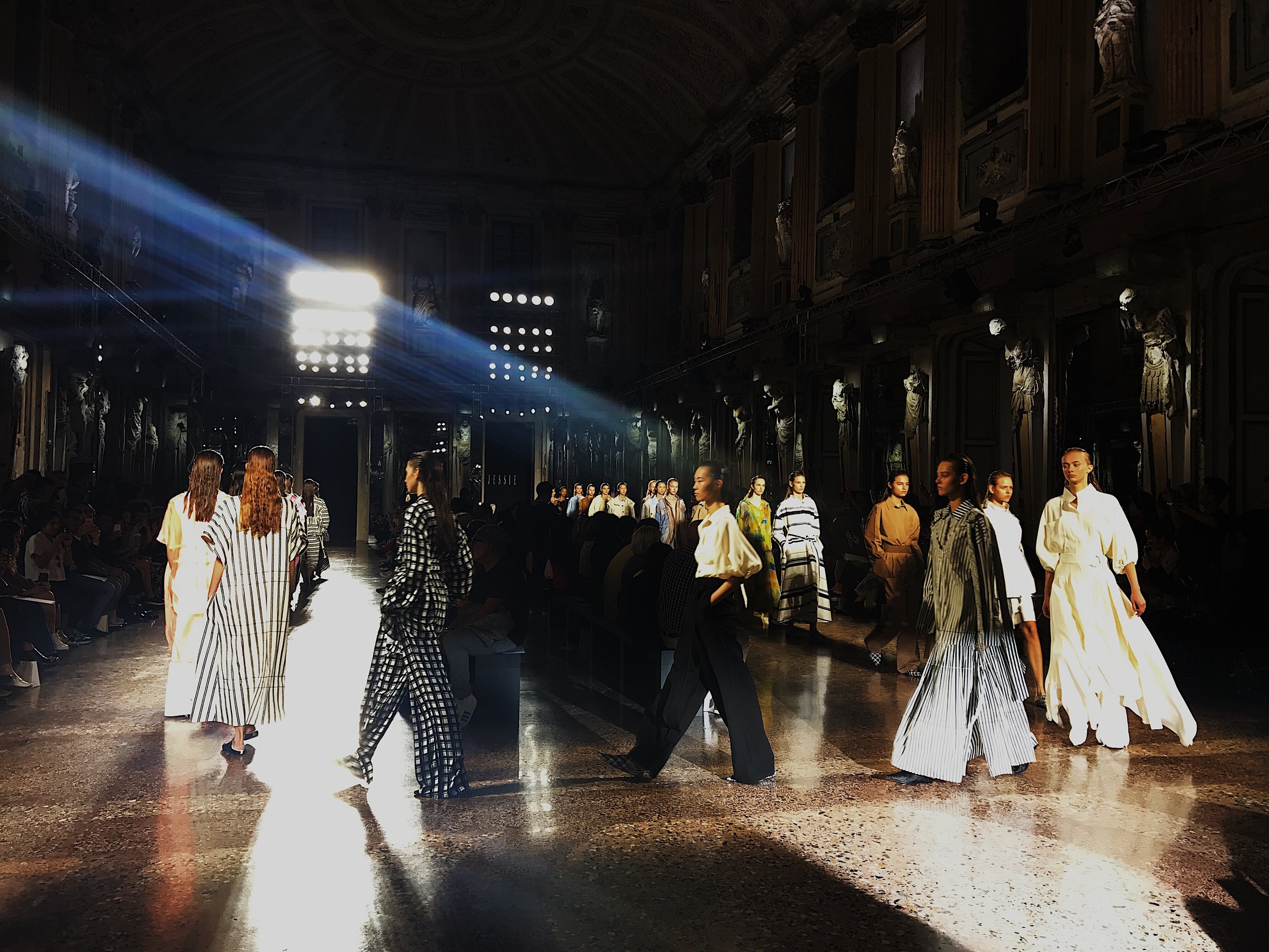 Models walking on runway during fashion show inside a grand hall with ornate walls and statues, illuminated by spotlight.