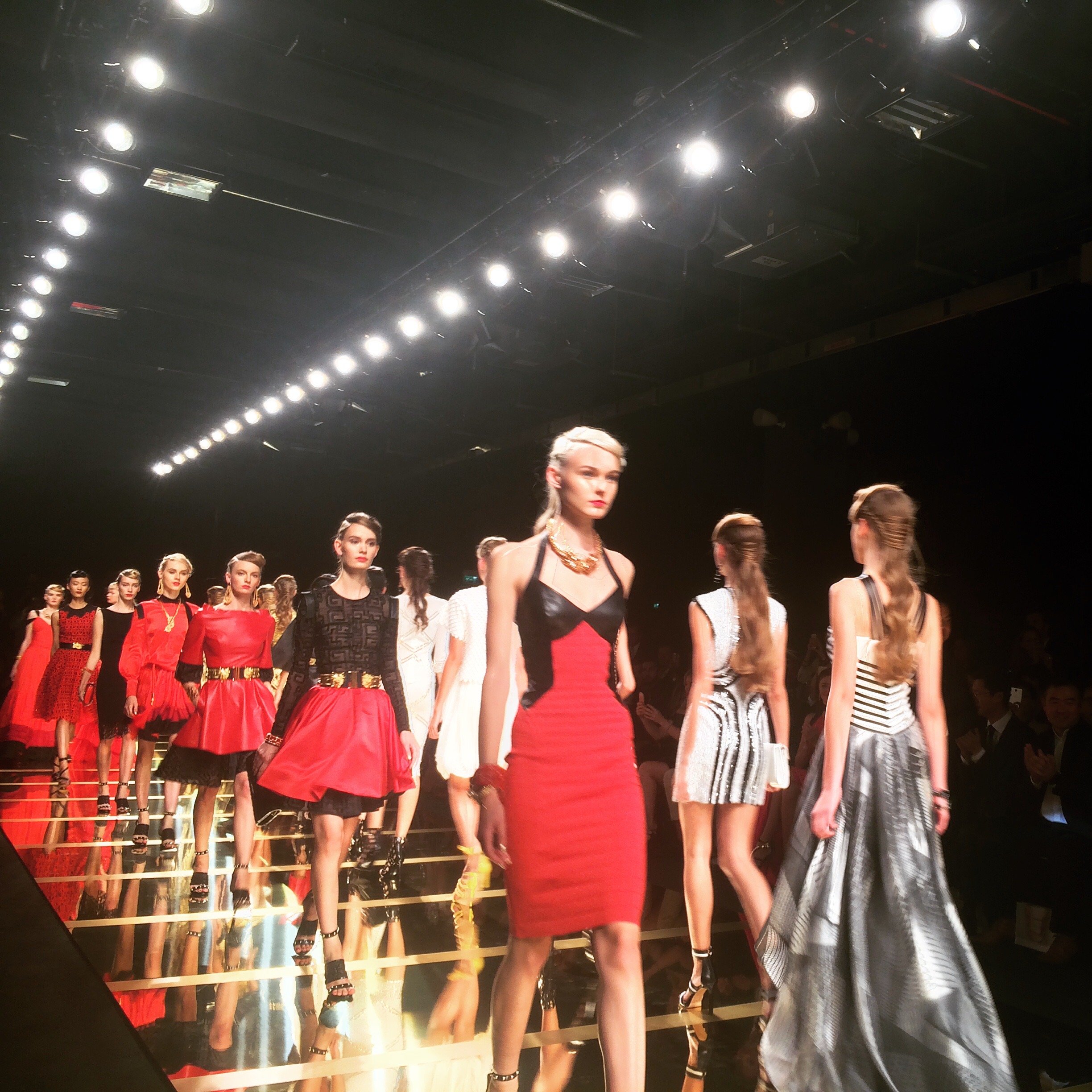 Models walking a runway during a fashion show, featuring models in various colorful and stylish outfits under bright stage lights.