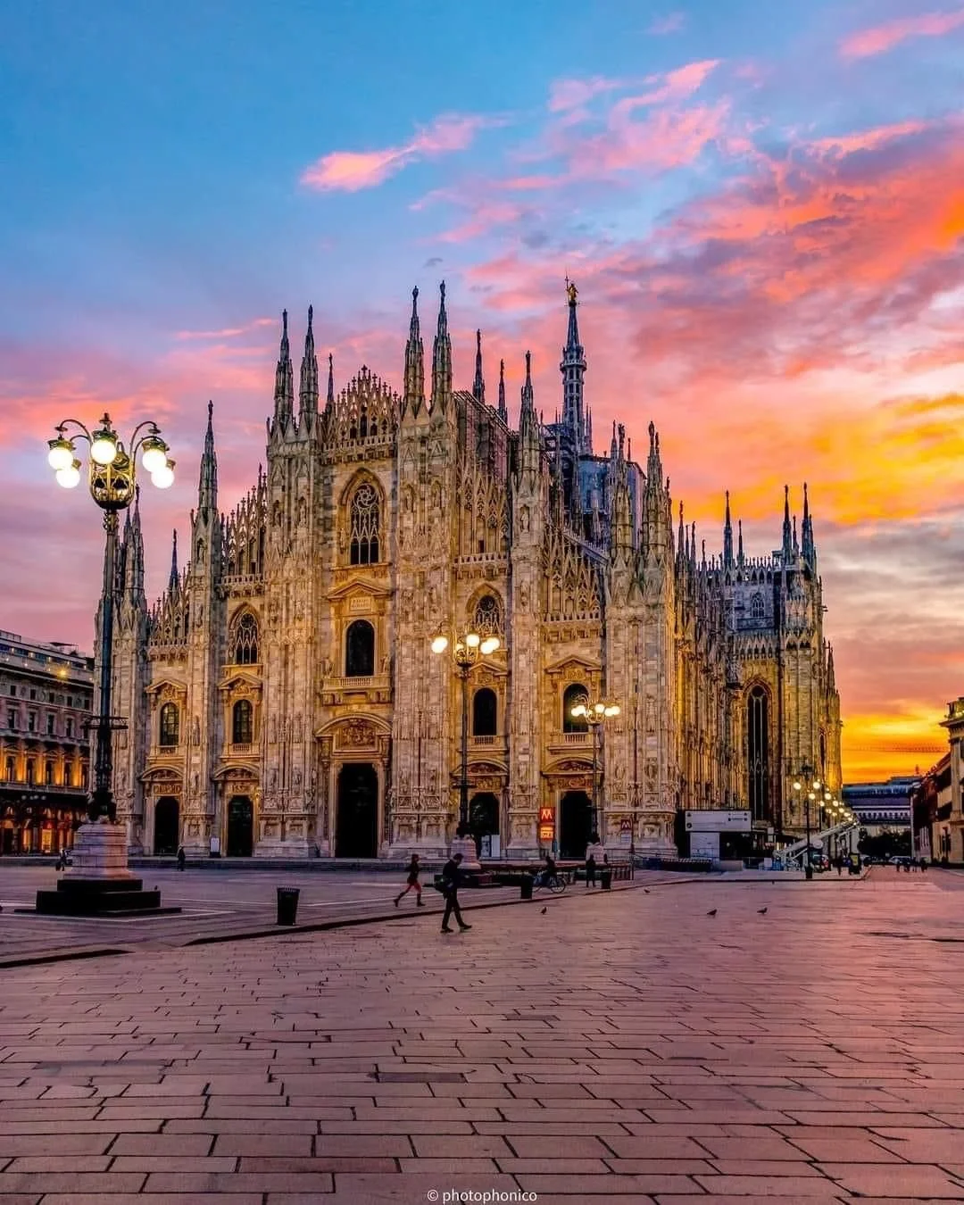 Sunset sky over the Milan Cathedral, a large Gothic cathedral with intricate spires and ornate details, located in an open plaza with few pedestrians and street lamps.