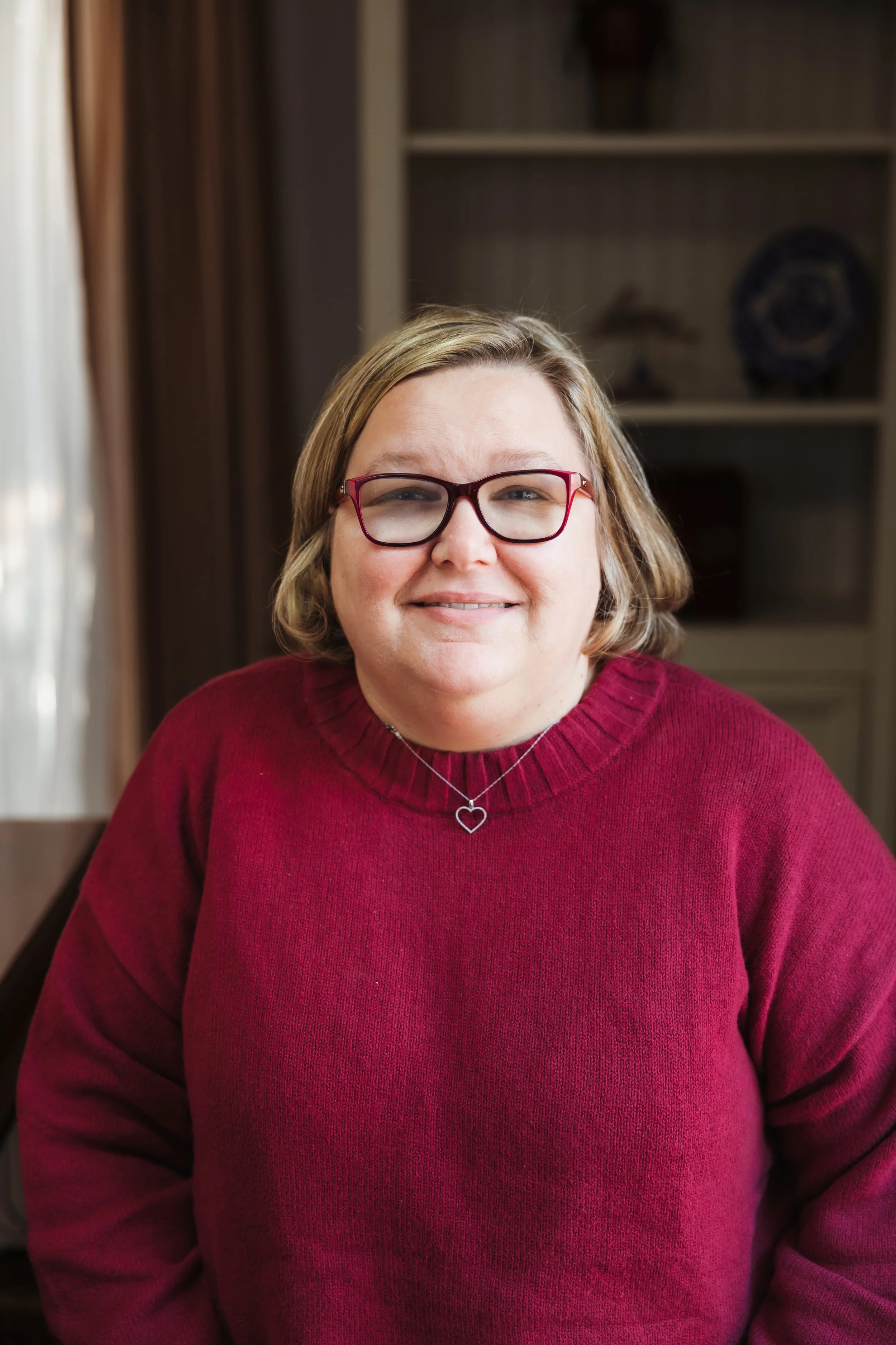 A woman with shoulder-length blonde hair and glasses, wearing a red sweater and heart-shaped necklace, smiling indoors with a bookshelf and a window in the background.