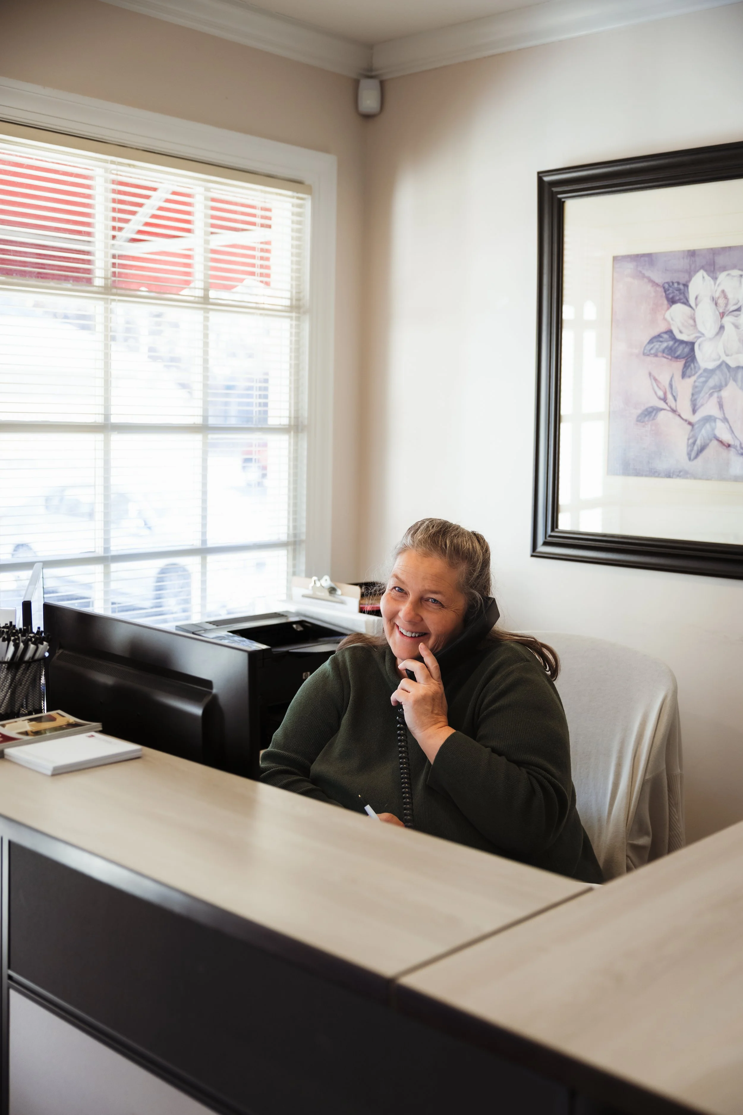 A woman sitting at a reception desk, smiling and talking on the phone in an office with a large window and framed floral artwork on the wall.