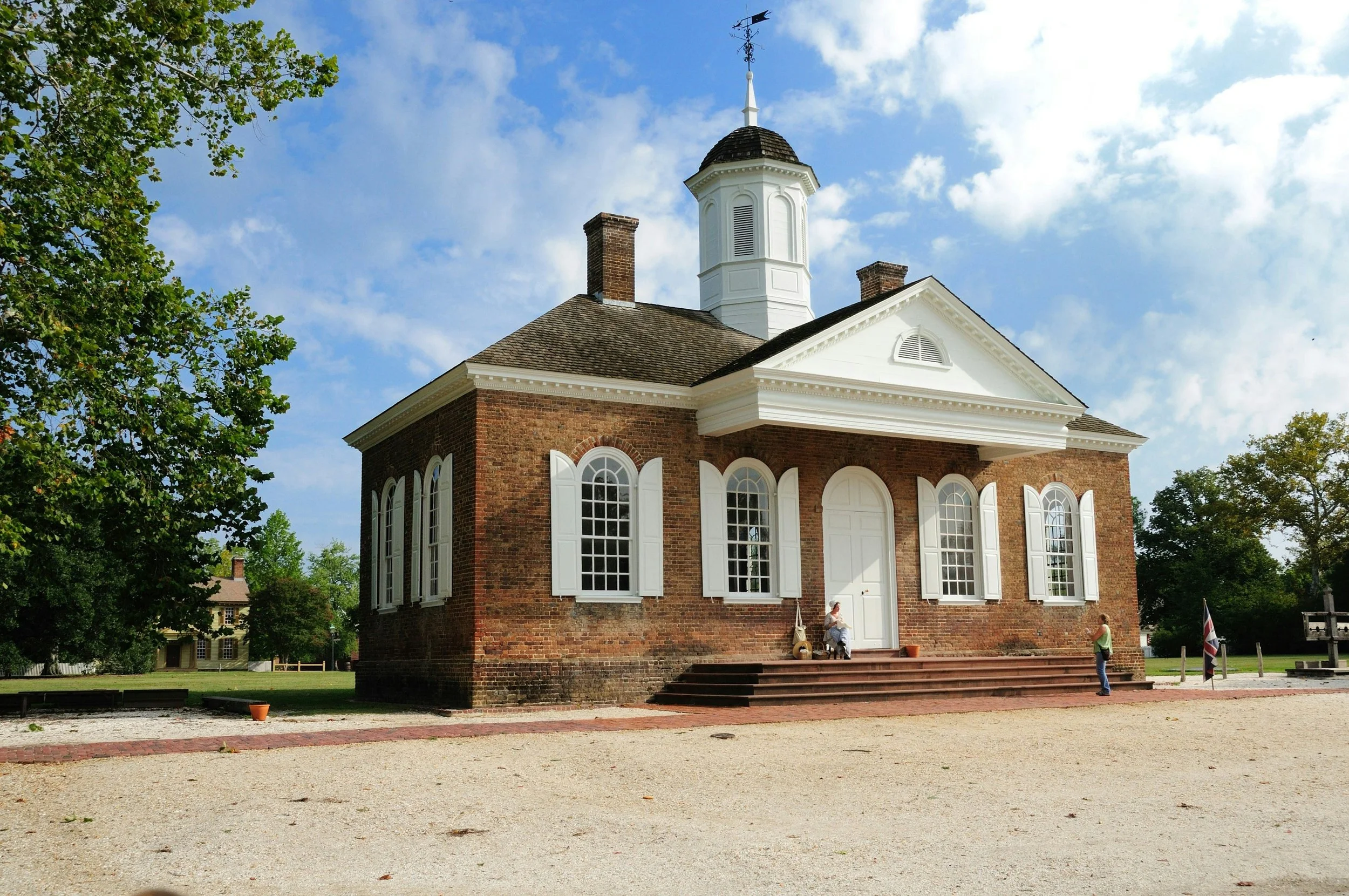 A historic brick church with white window shutters and trim, a white steeple, and stairs leading to a central door. Two women are seated on the steps, and another woman stands nearby. The church is surrounded by trees and a gravel yard, with a blue sky and clouds overhead.