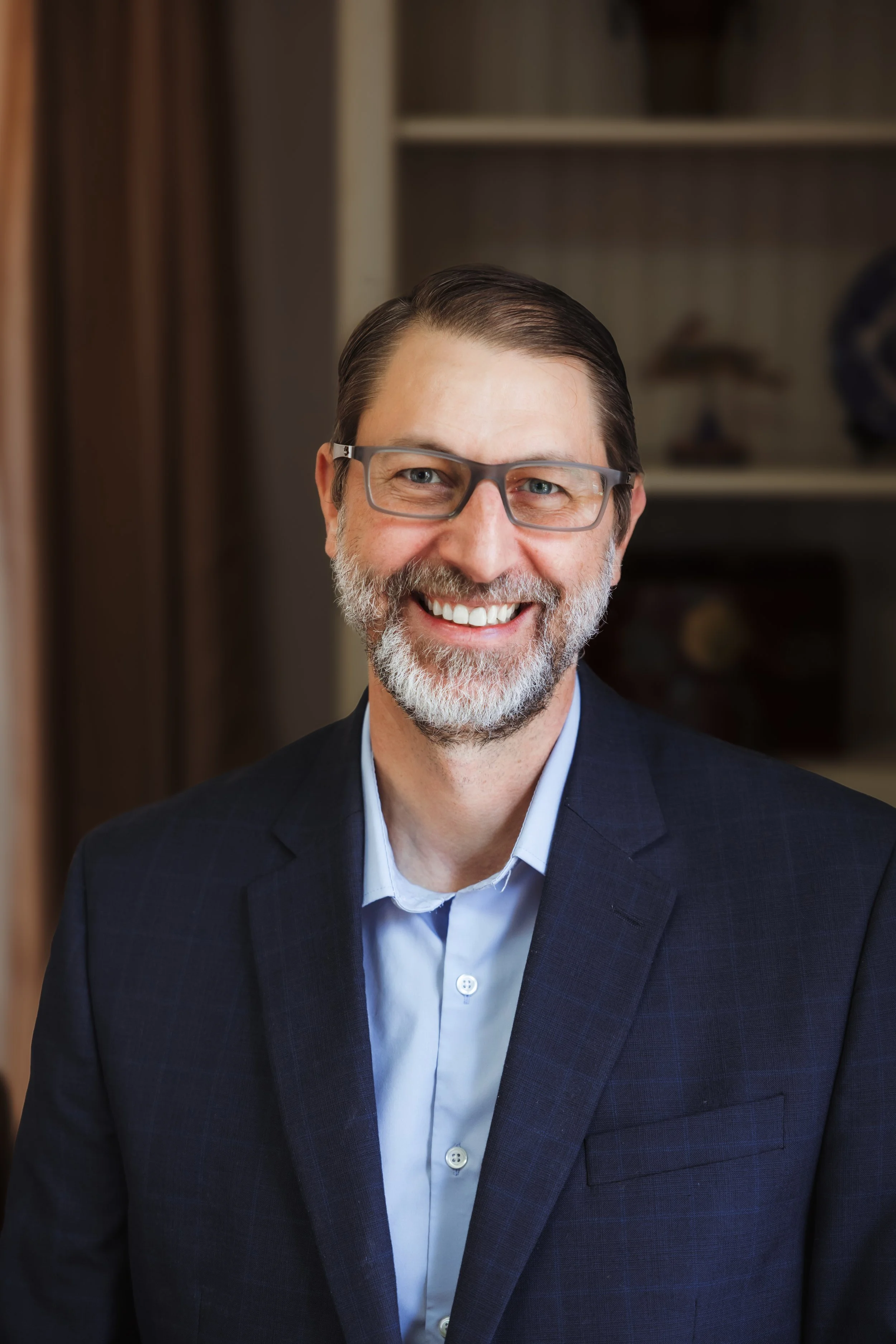 A smiling middle-aged man with glasses, a beard, and gray hair, wearing a dark suit jacket and light blue shirt, standing indoors against a blurred bookshelf background.