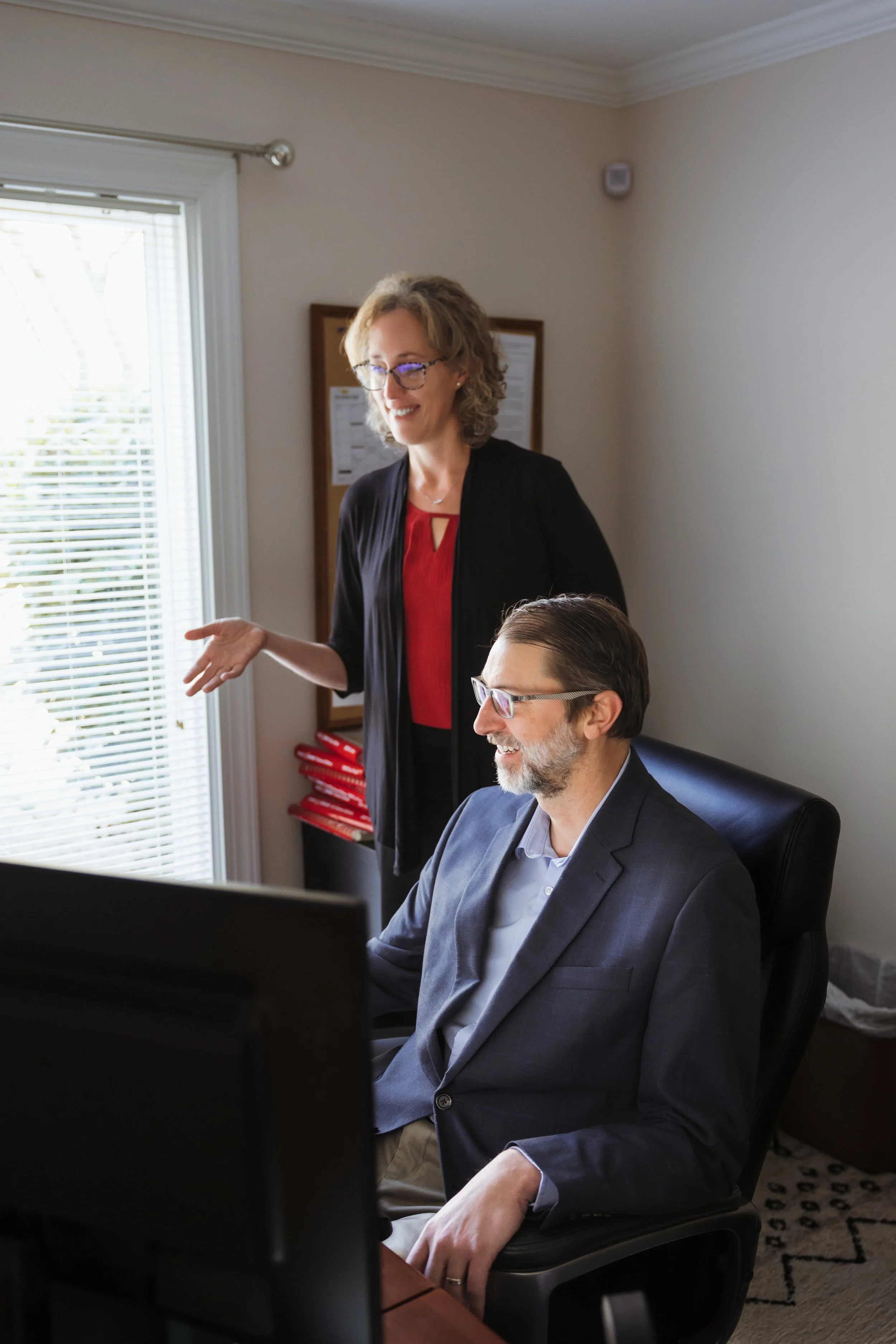 A woman standing and talking to a man sitting at a desk, working on a computer in an office with a window and blinds.