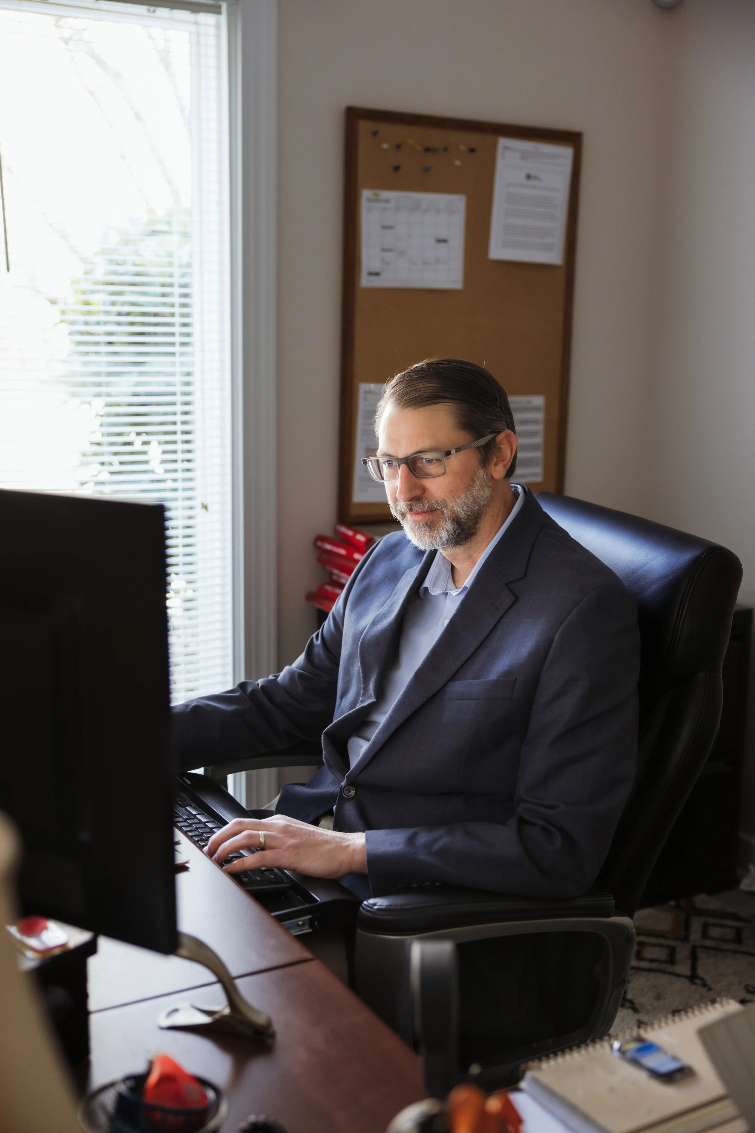 A man in a suit working on a computer in an office, with a corkboard and window in the background.