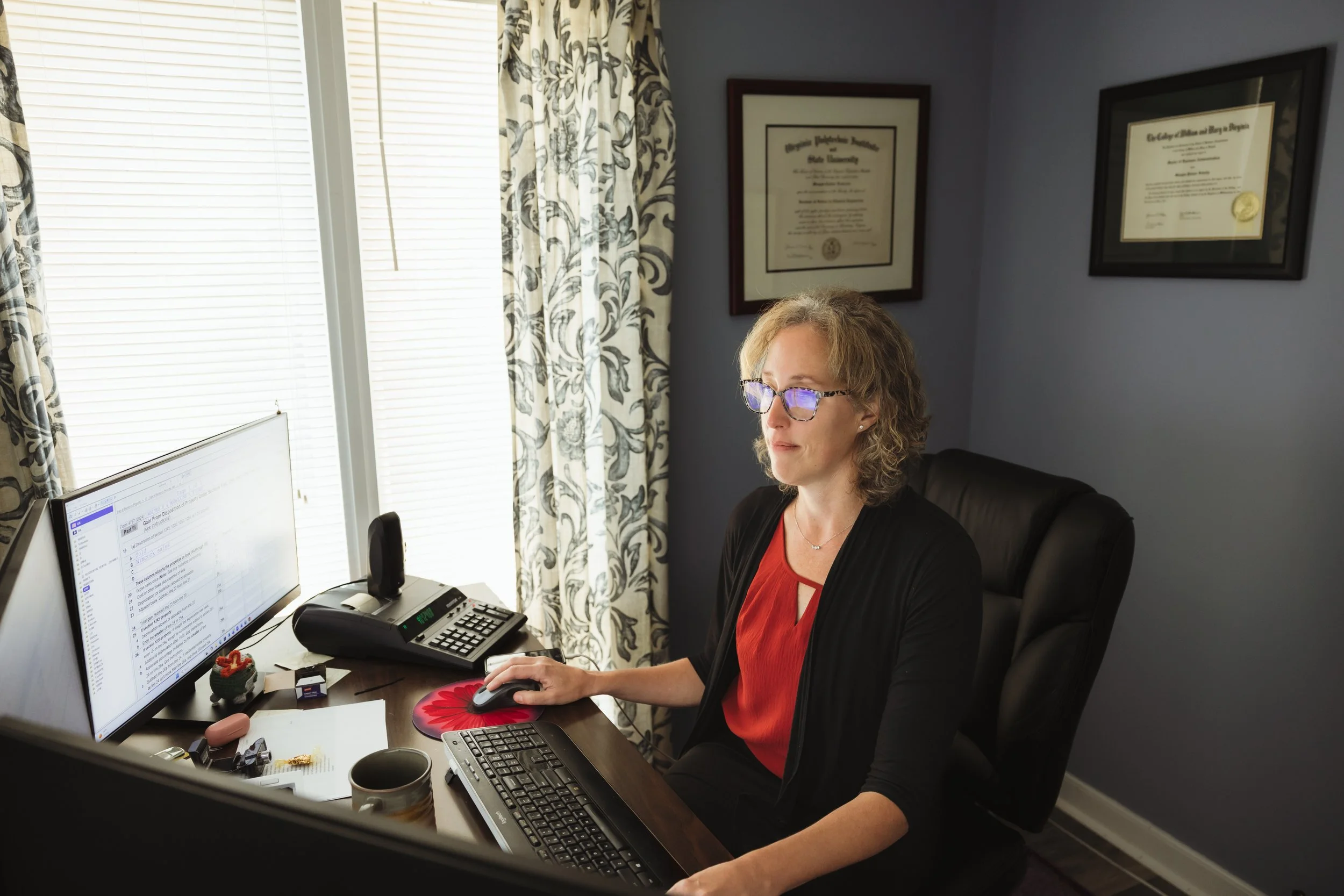 A woman with glasses sitting at a desk working on a computer, with diplomas hanging on the wall behind her.