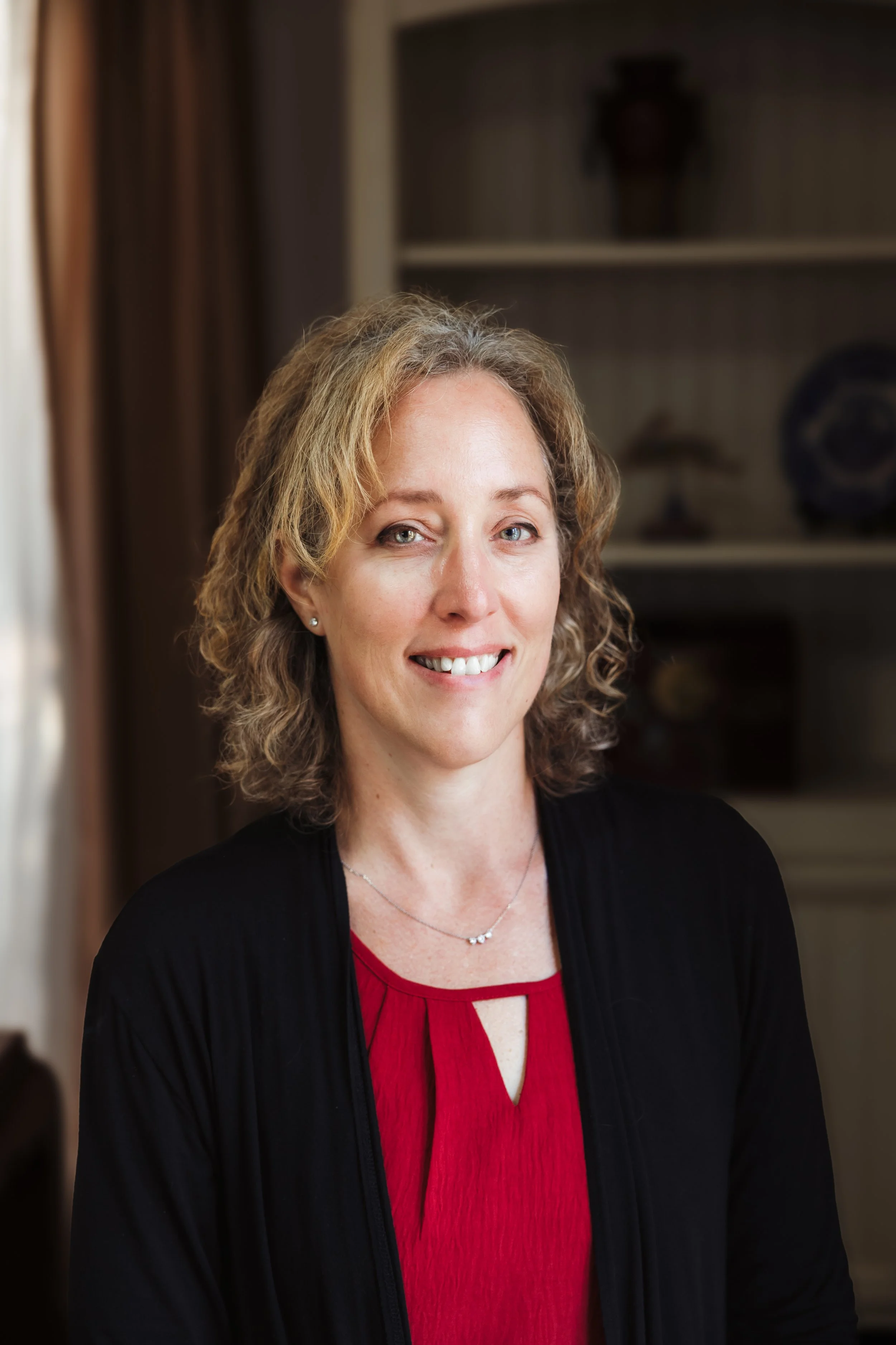 A woman with wavy blonde hair wearing a red blouse and black cardigan, smiling at the camera in a cozy home setting with a bookshelf in the background.