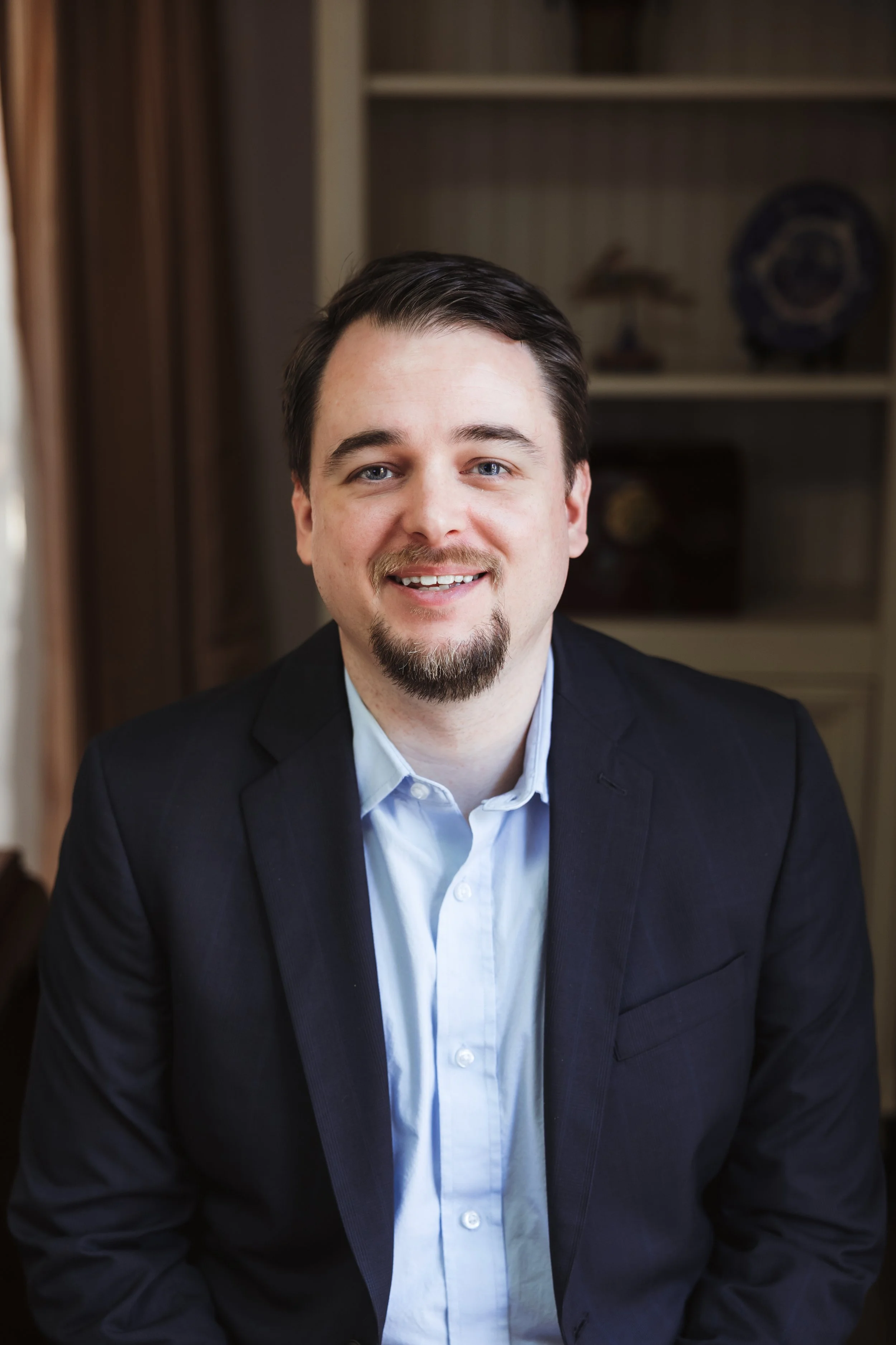 A man with light skin, dark brown hair, a goatee, and blue eyes wearing a dark blazer and light blue shirt, sitting indoors with a bookshelf in the background.