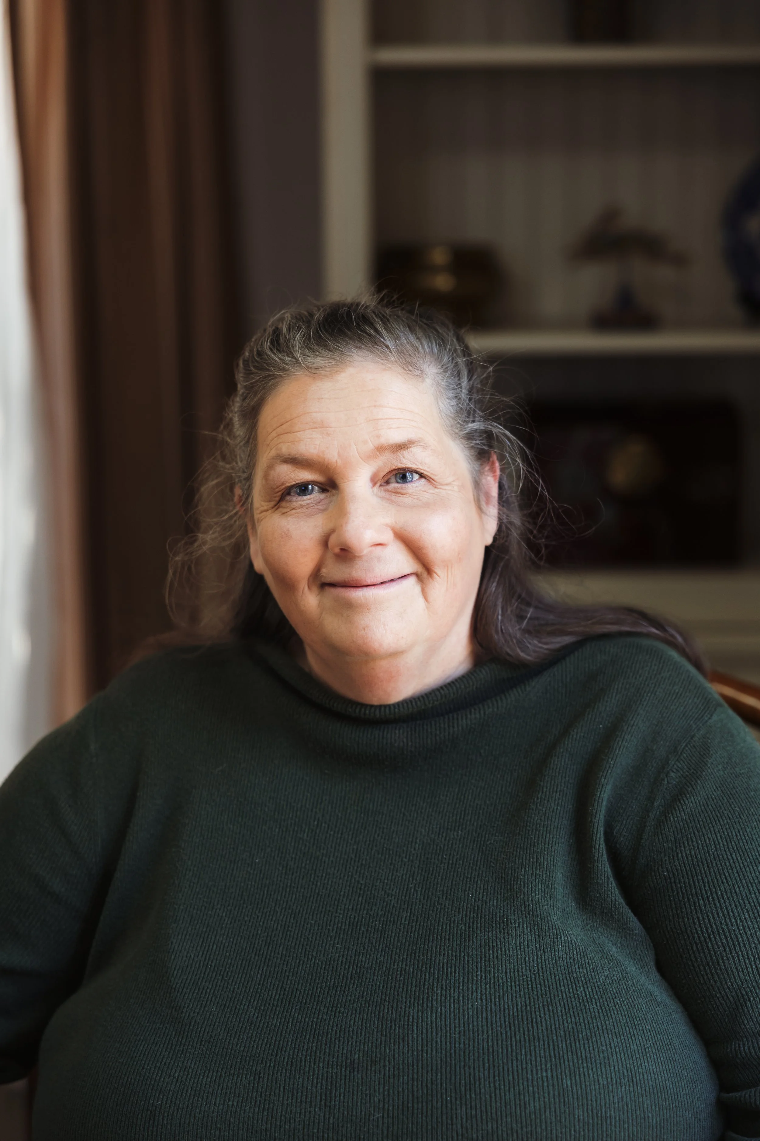 A middle-aged woman with long dark hair, wearing a dark green sweater, smiling while sitting indoors with a blurred background of shelves and decor.