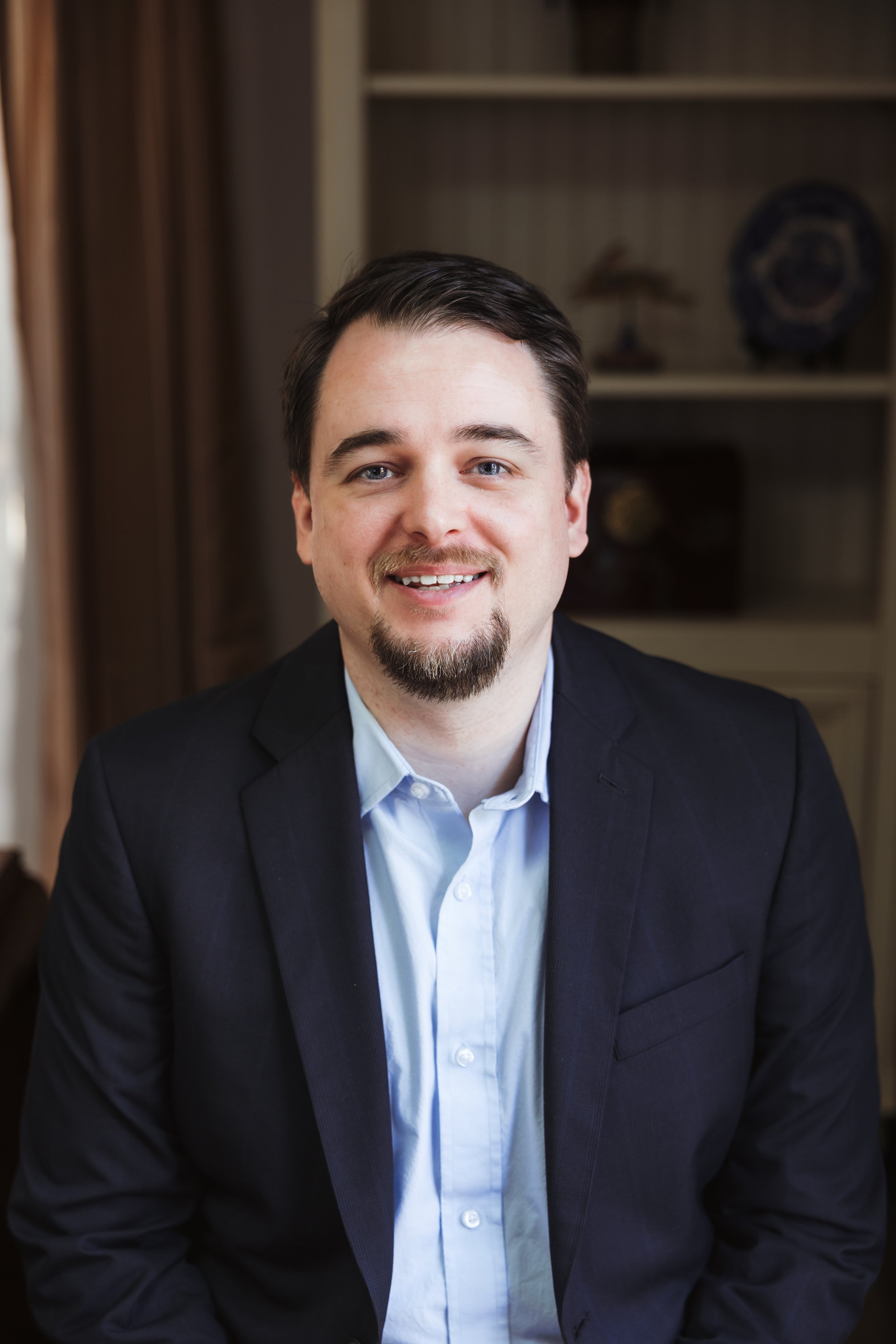 A man with dark hair, light skin, a goatee, wearing a white dress shirt and navy blazer, smiling in front of a blurred bookshelf background.