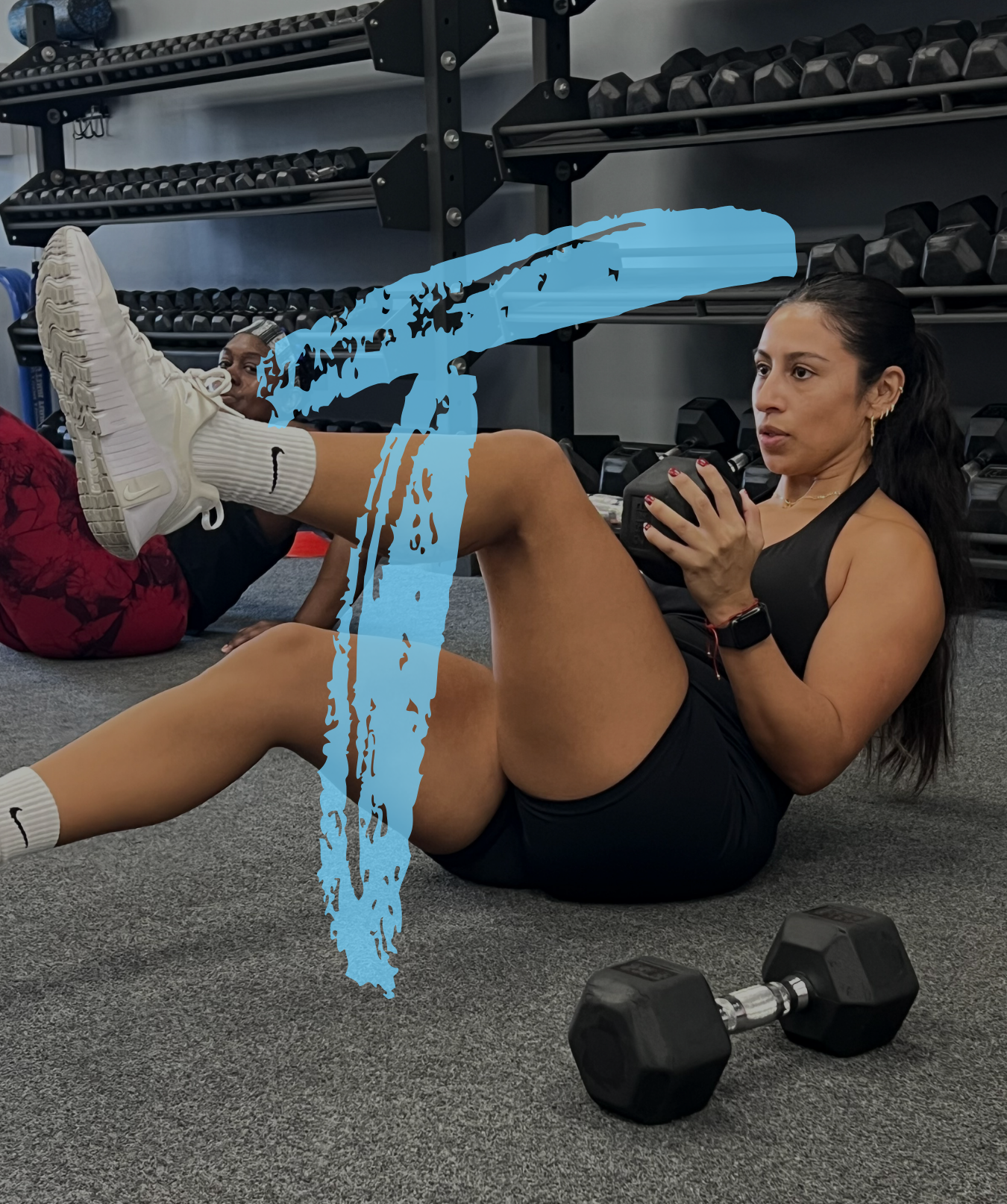 A woman in workout attire doing a core exercise on the gym floor with a dumbbell nearby, while another woman lies behind her during a workout session in a gym.