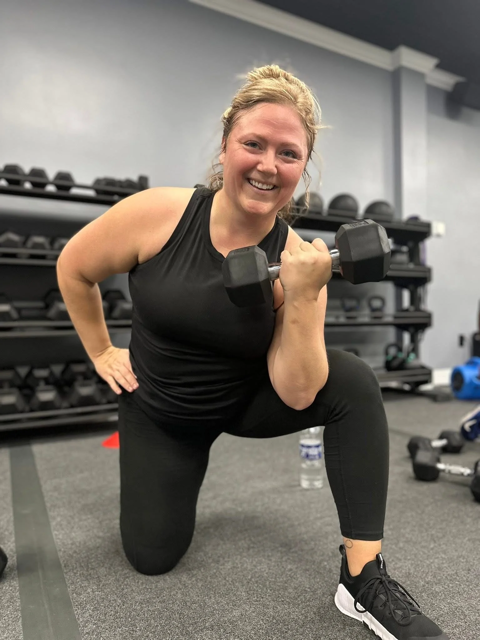 A woman in black workout clothes holding a dumbbell, kneeling in a gym with exercise equipment in the background, smiling at the camera.