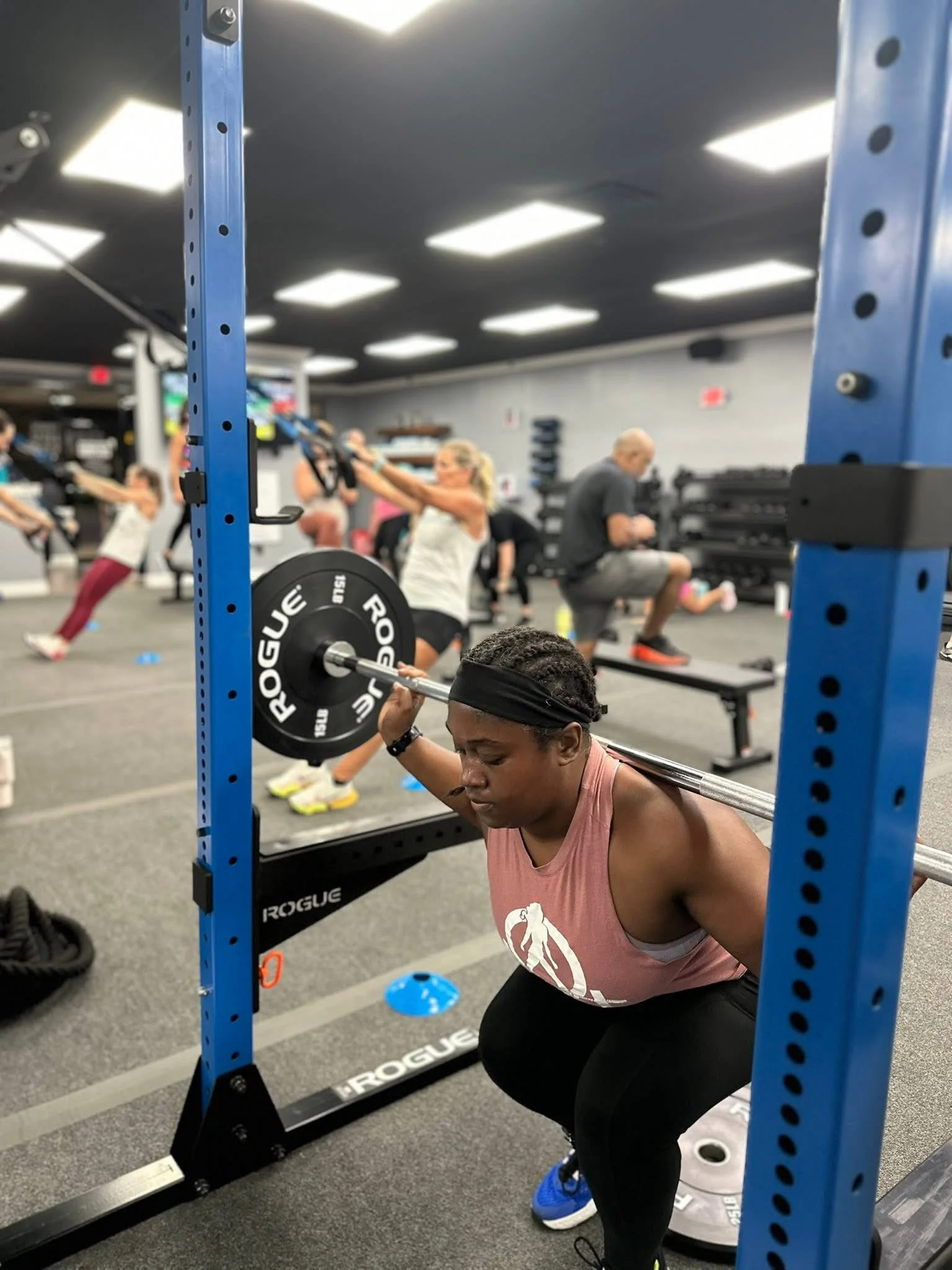 A woman performing a back squat exercise with a barbell loaded with weights at a gym, with other people exercising in the background.