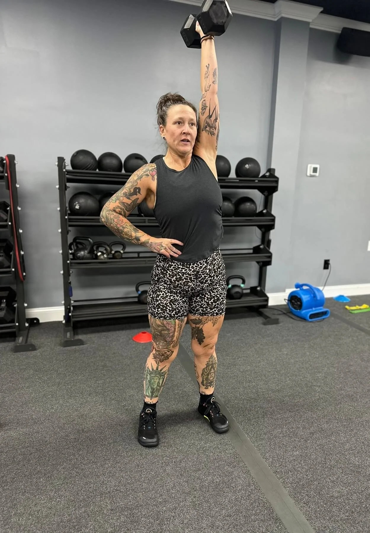A woman with tattoos wearing a black sleeveless top and animal print shorts exercises in a gym. She is holding a dumbbell overhead with her right arm, standing on a gray carpeted floor with gym equipment and black medicine balls in the background.