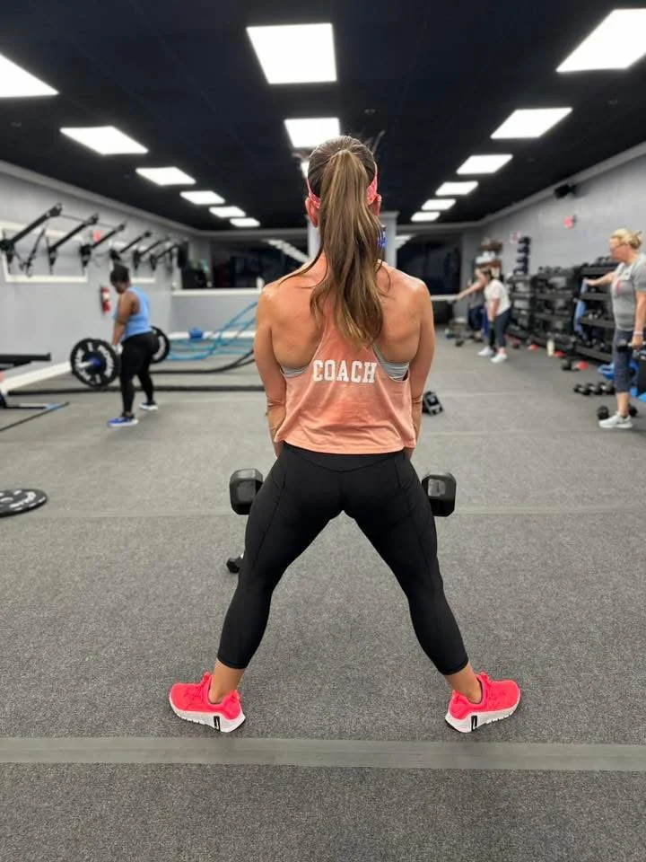 A woman with long hair in a ponytail, wearing a pink sleeveless top with the word 'COACH' on the back, black leggings, and pink sneakers, is standing in a gym facing away from the camera and holding dumbbells at her sides.